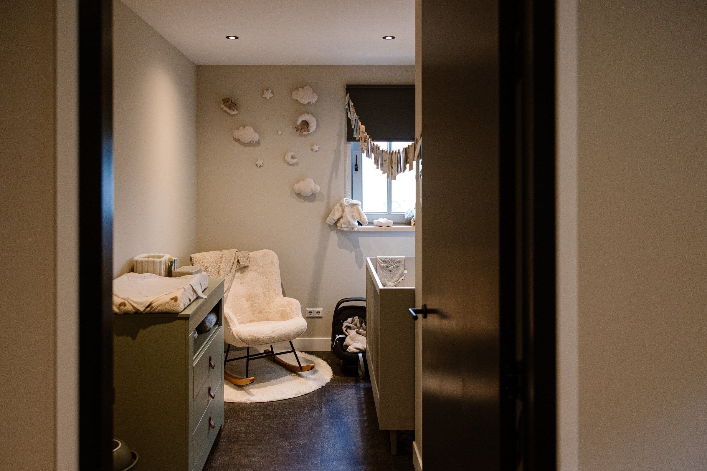 Doorkijk naar een babykamer met een zachte schommelstoel, een commode links, een babybed rechts en wolk- en sterrenmuurdecoratie bij het raam.