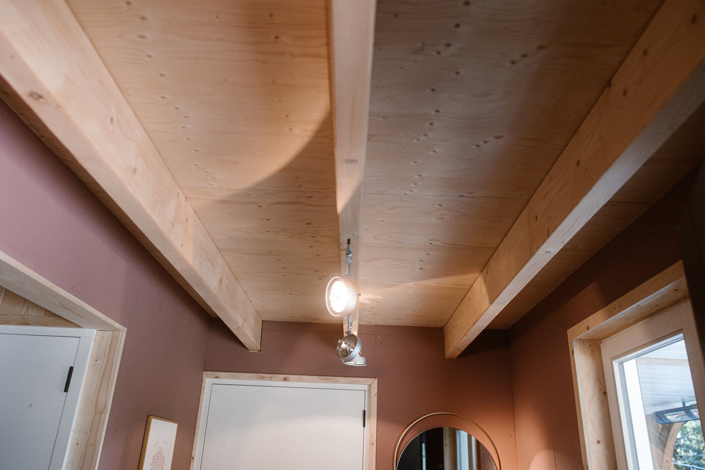 Kamer met houten plafonplaten en balken, paarsbruine muren, een witte deur en een ronde spiegel.