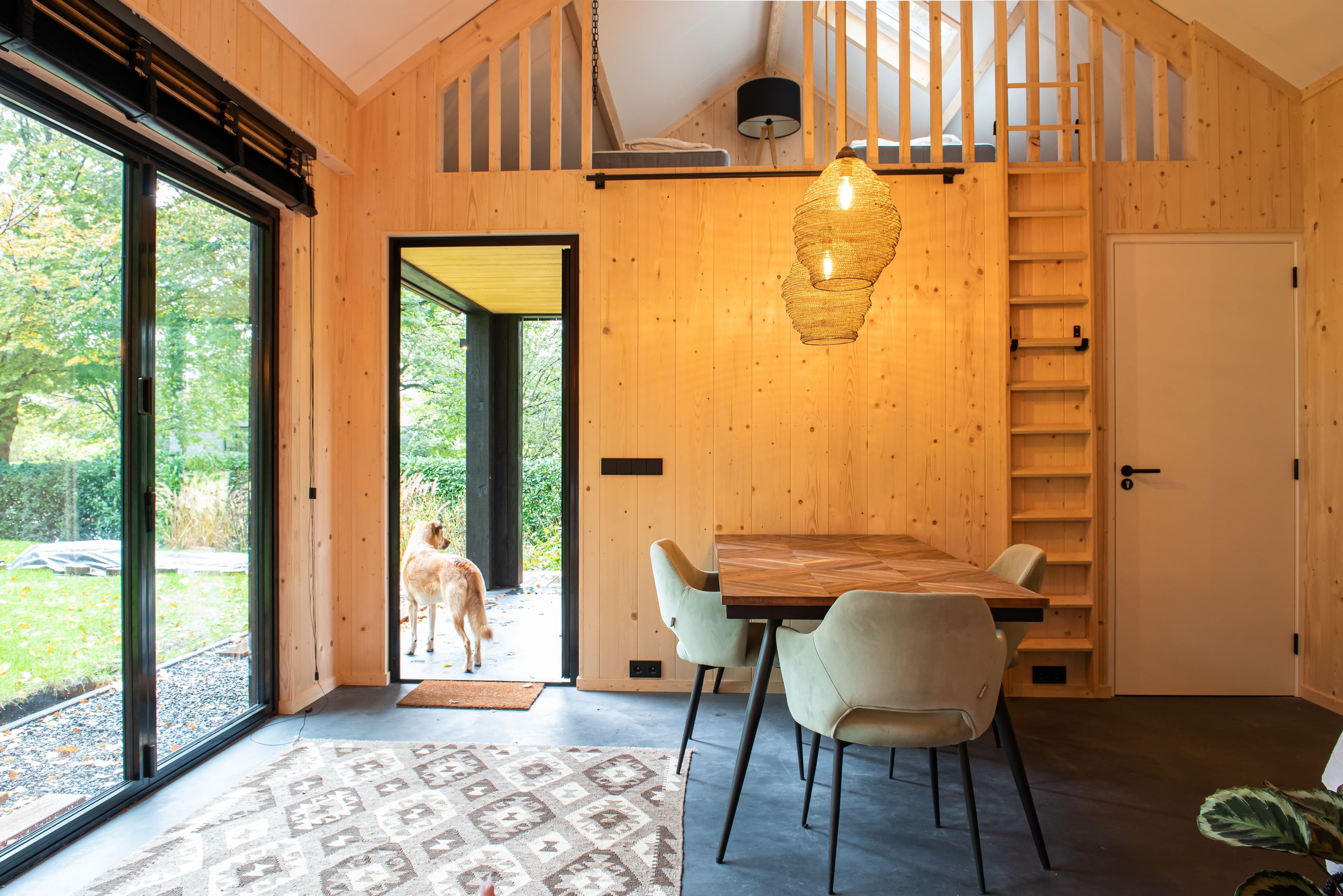 Binnenruimte van een huis met houten wanden, een houten eettafel met stoelen, een open deur waar een hond buiten staat, en een trap naar een zolderruimte.