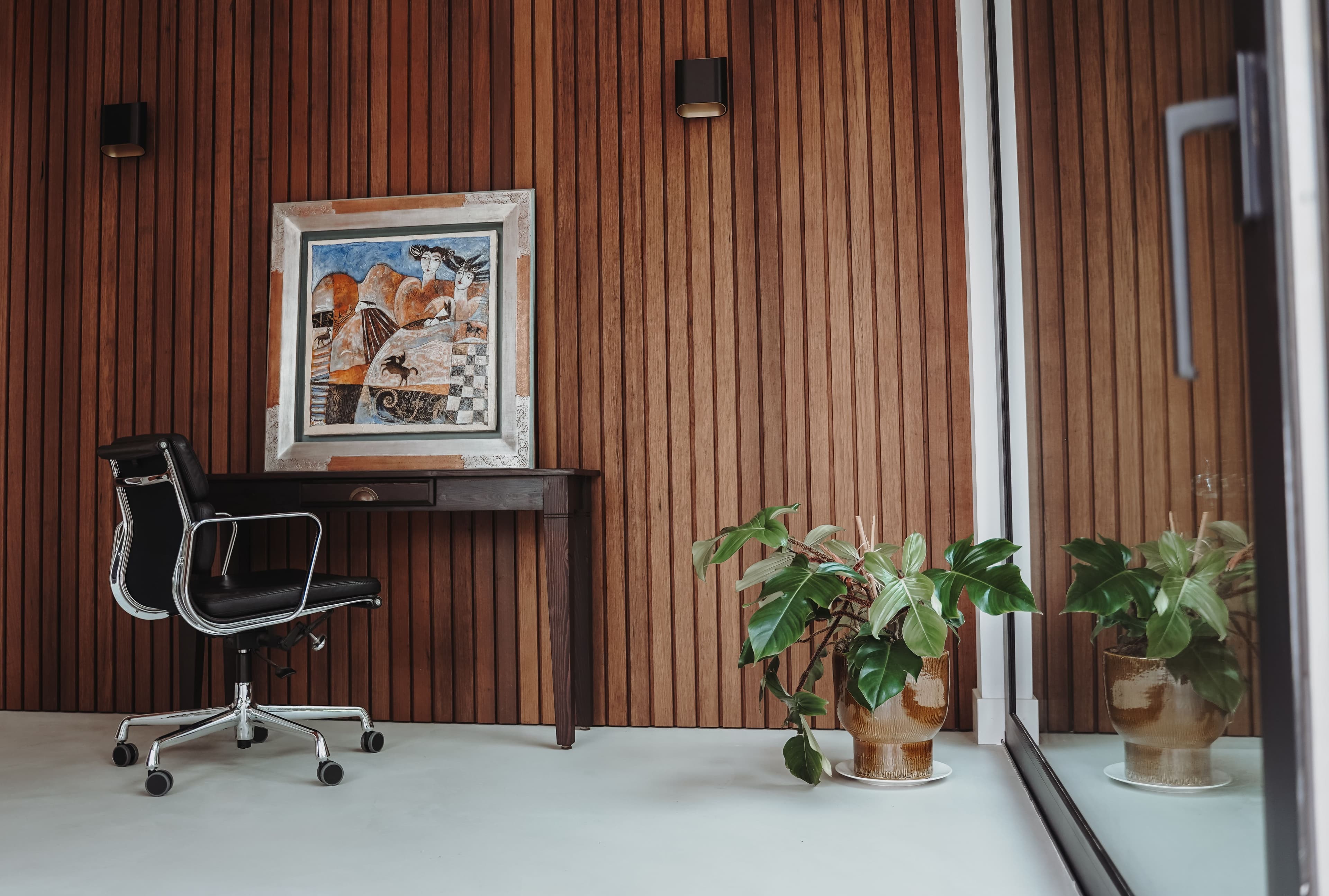 Kantoorruimte met houten lambrisering, een zwart bureaustoel, een bureau met schilderij en een plantenpot naast een glazen wand