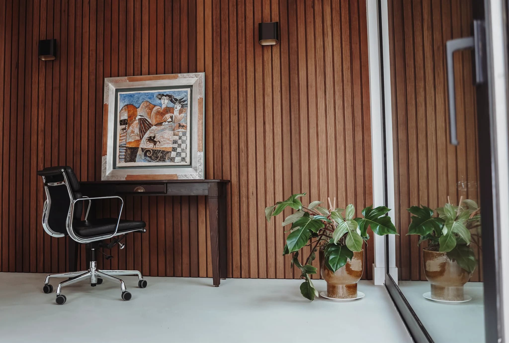 Kantoorruimte met houten lambrisering, een zwart bureaustoel, een bureau met schilderij en een plantenpot naast een glazen wand