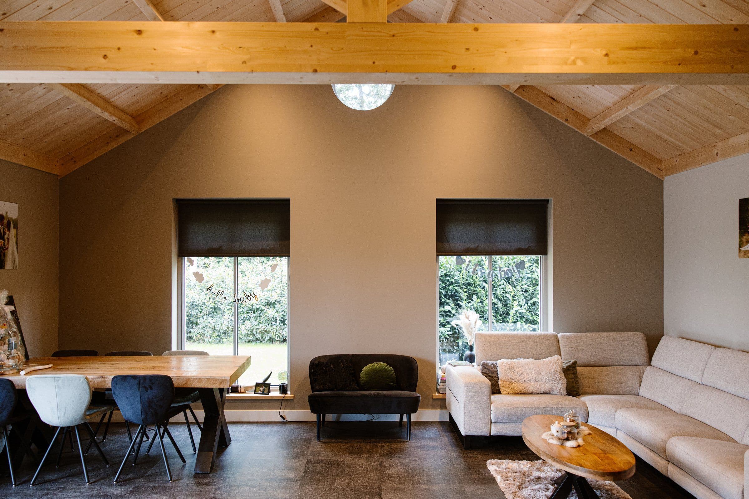Binnenruimte met houten plafond, eettafel met diverse stoelen, bank tussen ramen en hoekbank met salontafel.