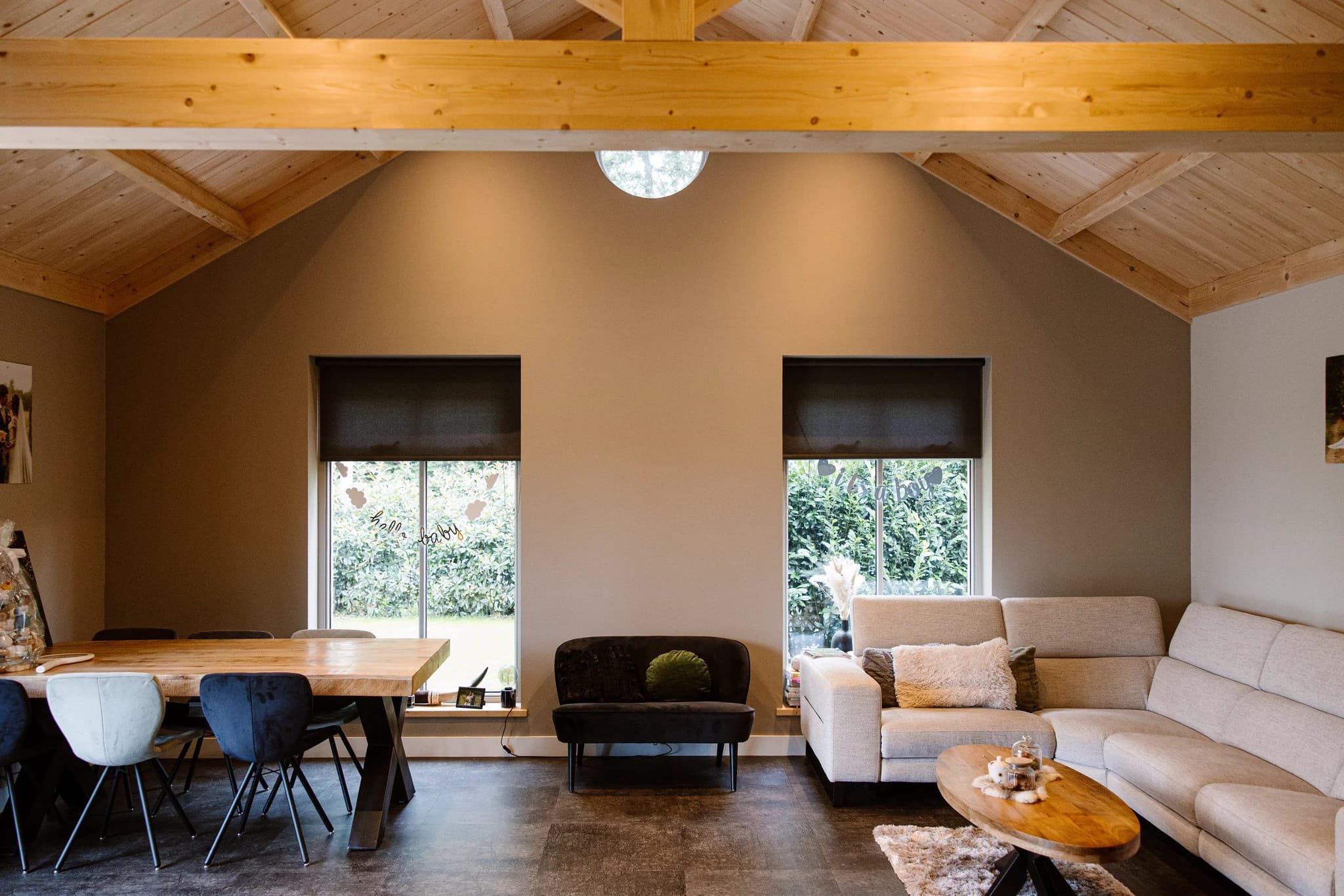 Binnenruimte met houten plafond, eettafel met diverse stoelen, bank tussen ramen en hoekbank met salontafel.