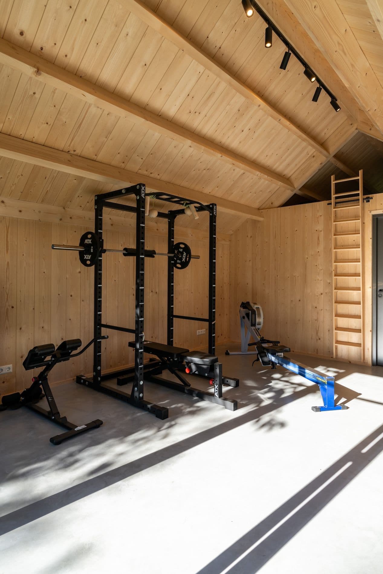 Binnenruimte met houten wanden en diversen fitnessapparaten, waaronder een bankdrukrek en roeimachine