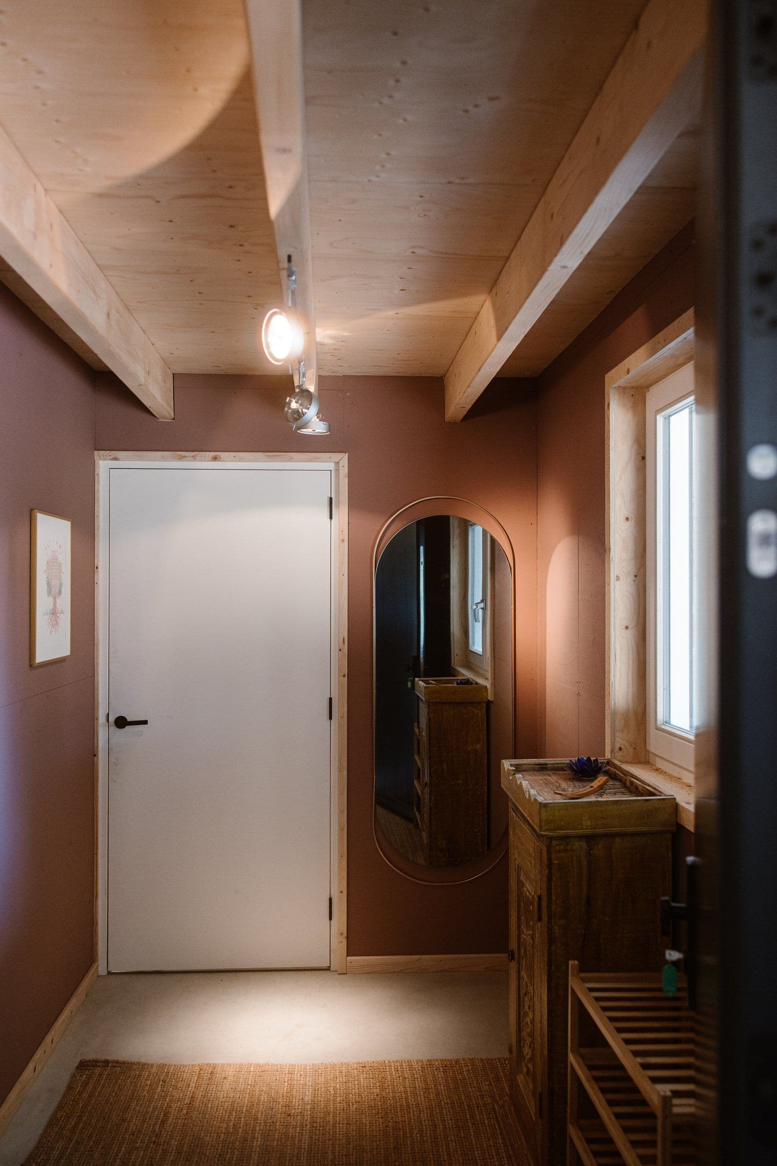 Binnenruimte met witte deur, ovale spiegel aan bruine muur, houten kastje, raam en houten balken aan het plafond