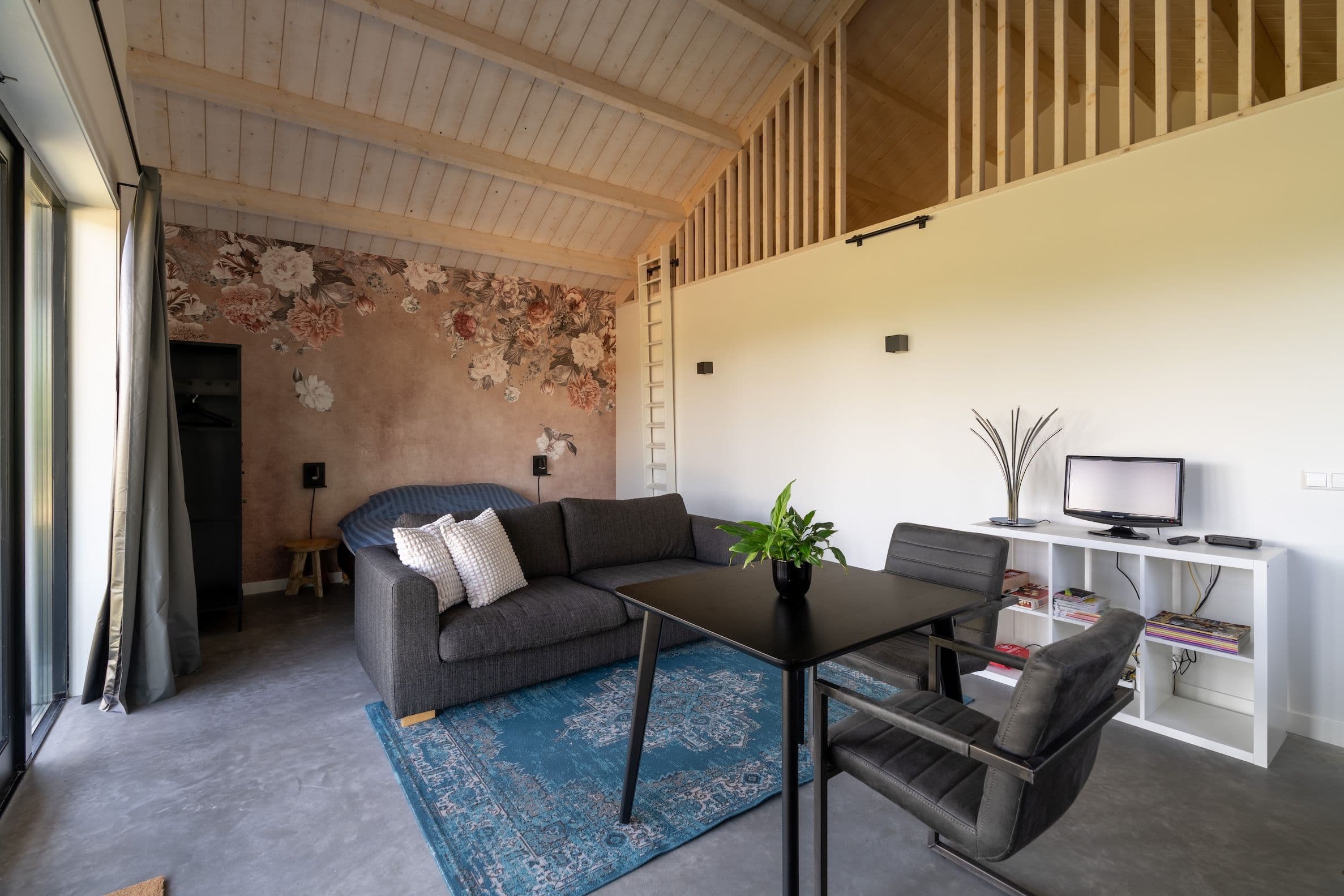 Gezellige binnenruimte met grijze bank, zwarte eettafel met stoelen, tv op witte kast en bed met bloemenmuur.