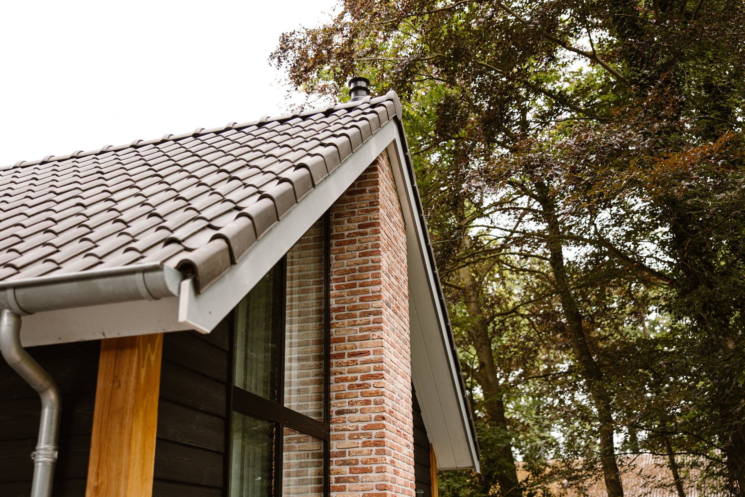 Dak met grijze dakpannen naast een bakstenen muur en zwarte houten gevel, met bomen op de achtergrond