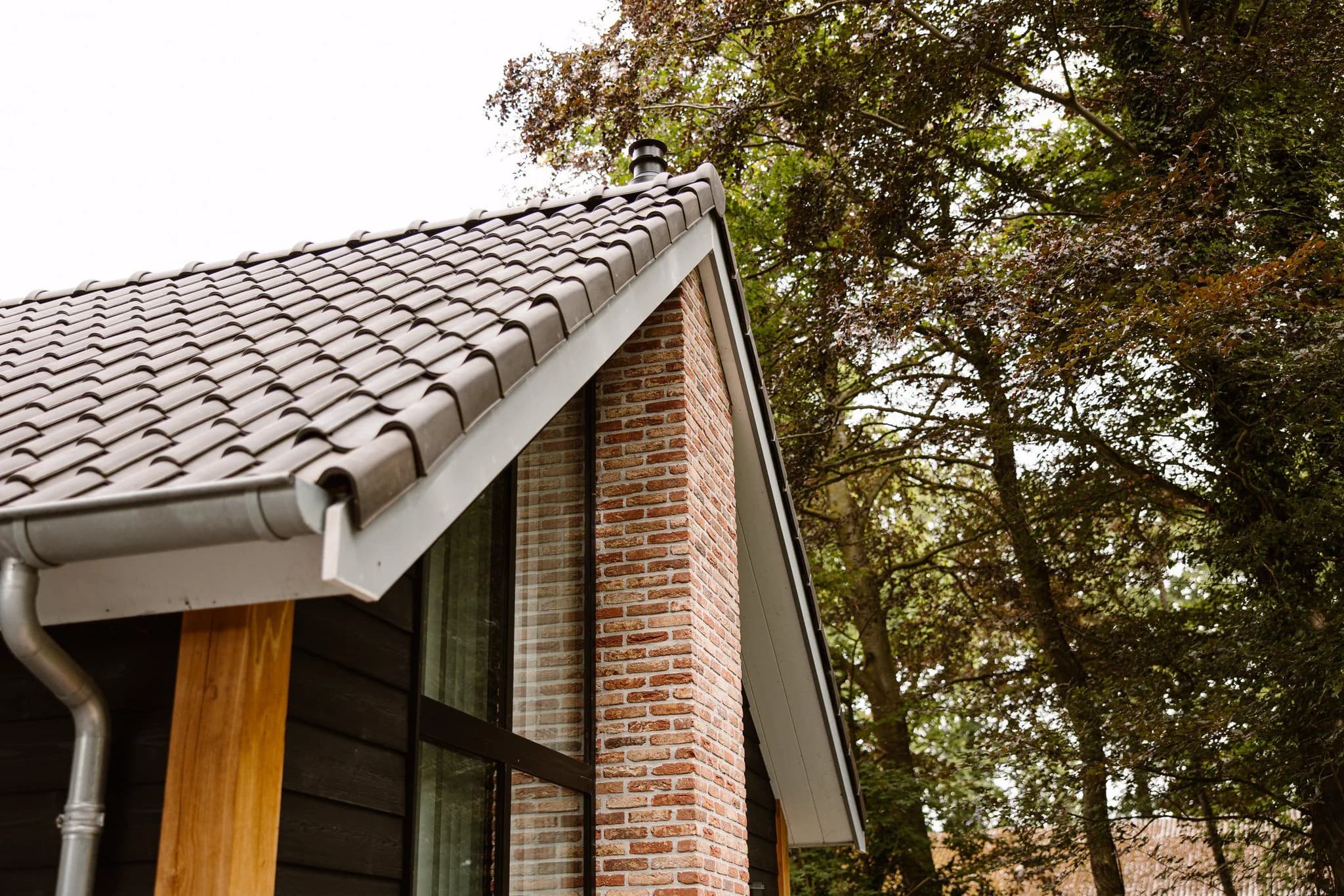 Dak met grijze dakpannen naast een bakstenen muur en zwarte houten gevel, met bomen op de achtergrond