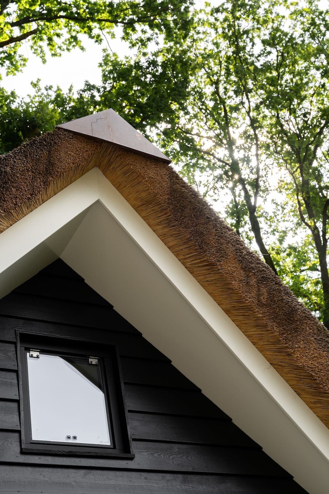Hoek van huis met rietdak, witte boeidelen en zwarte houten gevel met klein raam; bomen op de achtergrond
