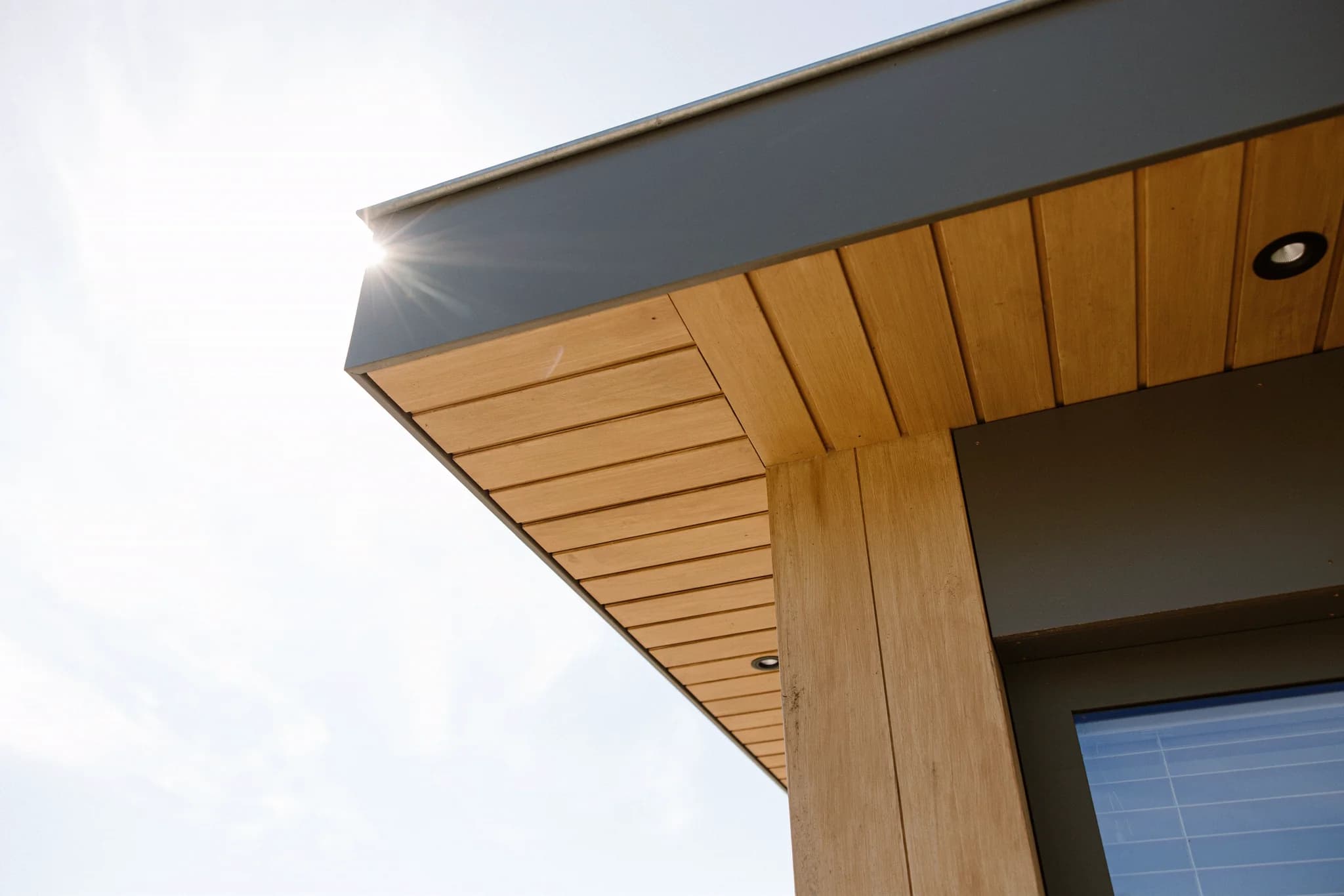 Hoek van een gebouw met houten bekleding en metalen dakrand tegen een blauwe lucht met zonlicht