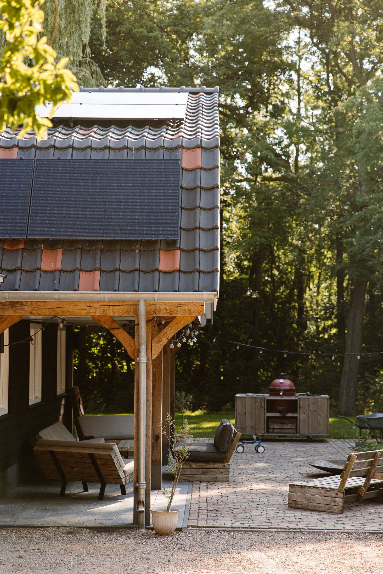 Veranda met houten banken en zonnepanelen op het dak, achtertuin met barbecue en bomen op de achtergrond