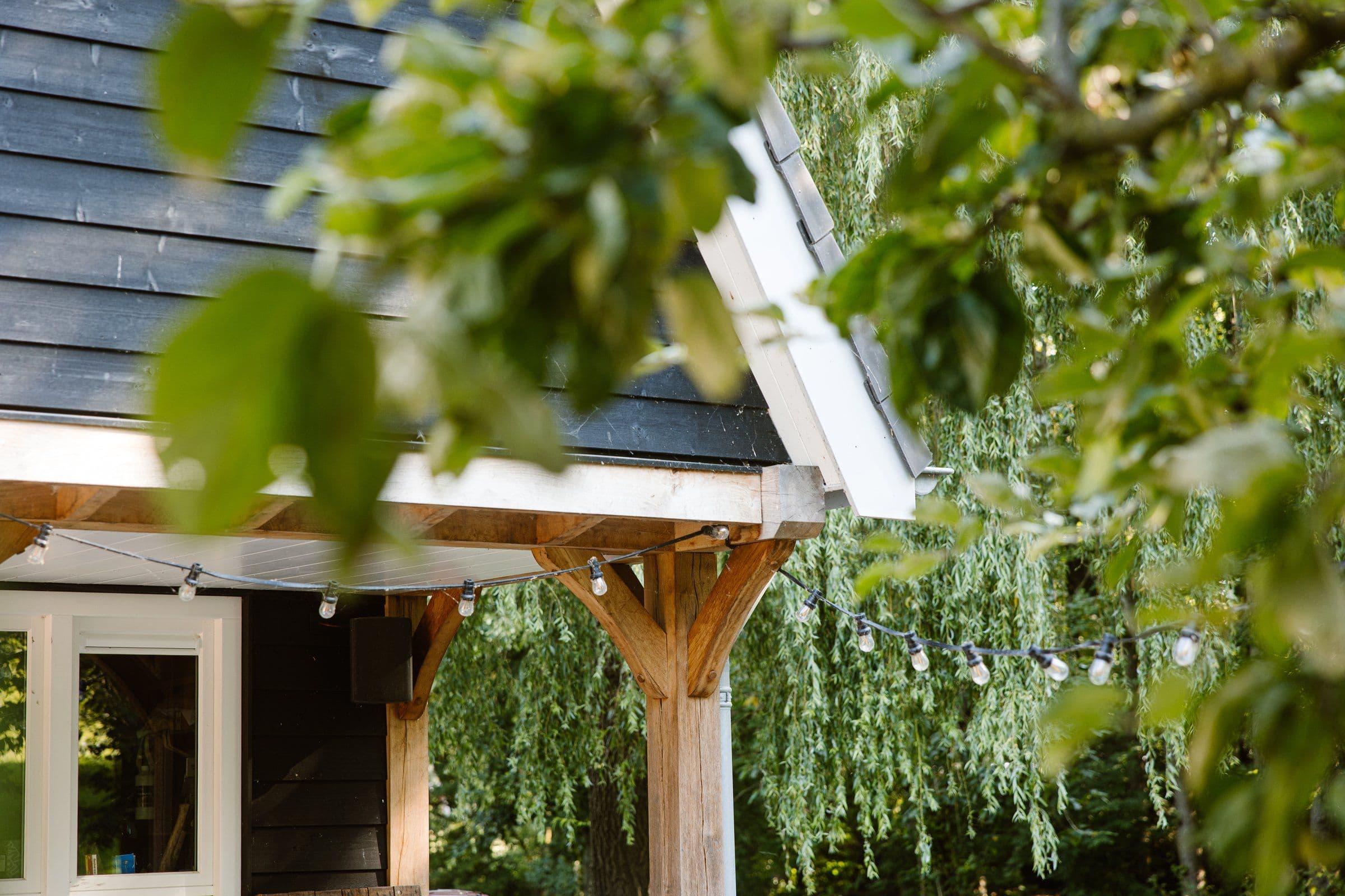 Houten veranda met zwarte en witte gevel, omgeven door groene bladeren en een slinger met lichtjes