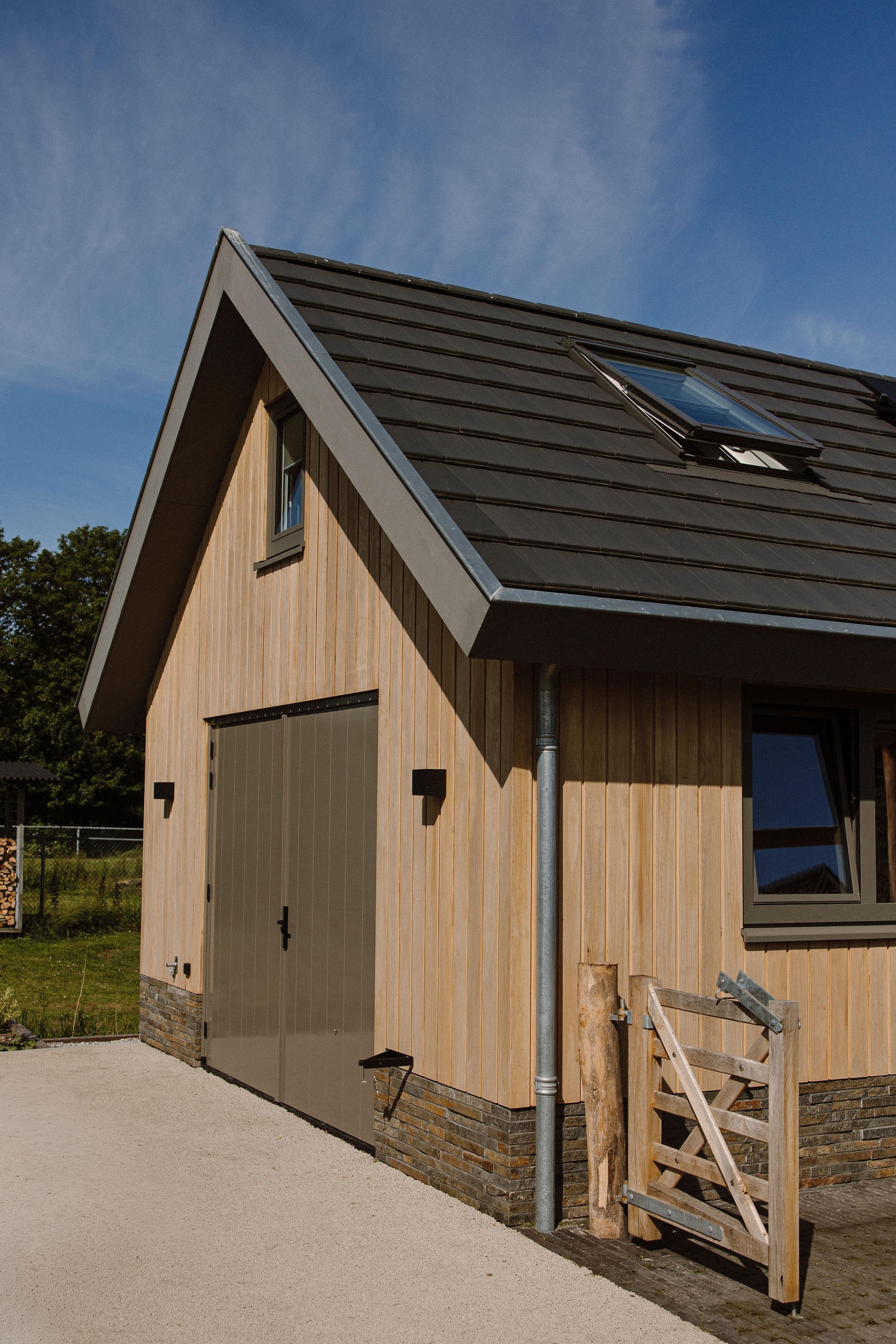 Zijaanzicht van een houten bijgebouw met verticale houten bekleding, donkere dakbedekking met dakraam en een gesloten garagedeur; blauwe lucht op de achtergrond.