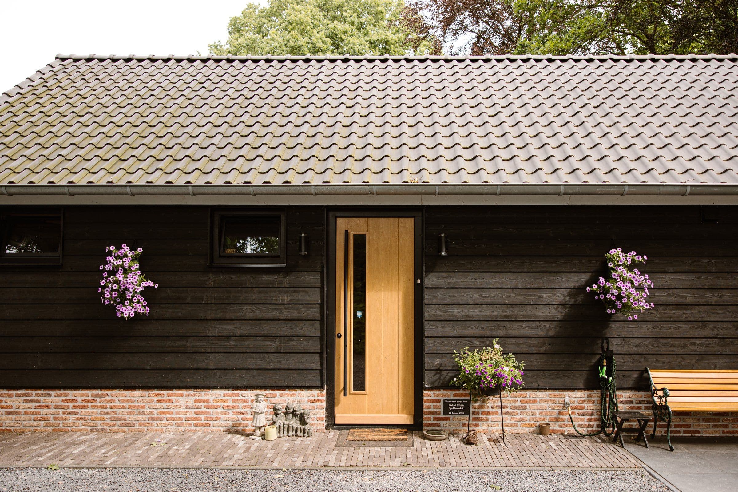 Vooraanzicht van een houten huis met een lichtbruine deur, kleine ramen, hangende roze bloemen, een houten bank en tuinbeeldjes naast de deur.