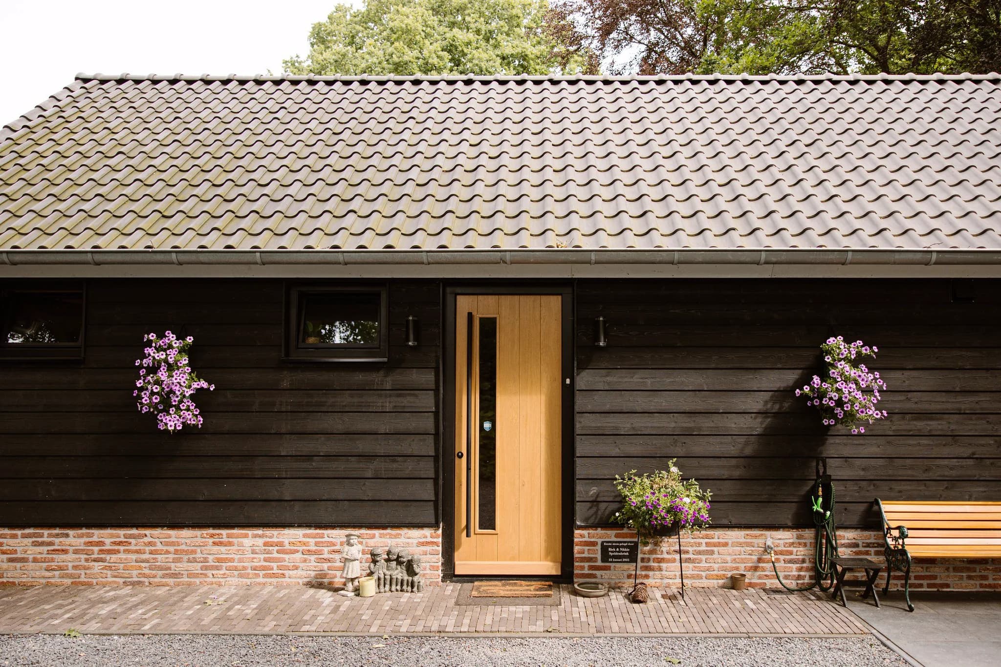 Vooraanzicht van een houten huis met een lichtbruine deur, kleine ramen, hangende roze bloemen, een houten bank en tuinbeeldjes naast de deur.