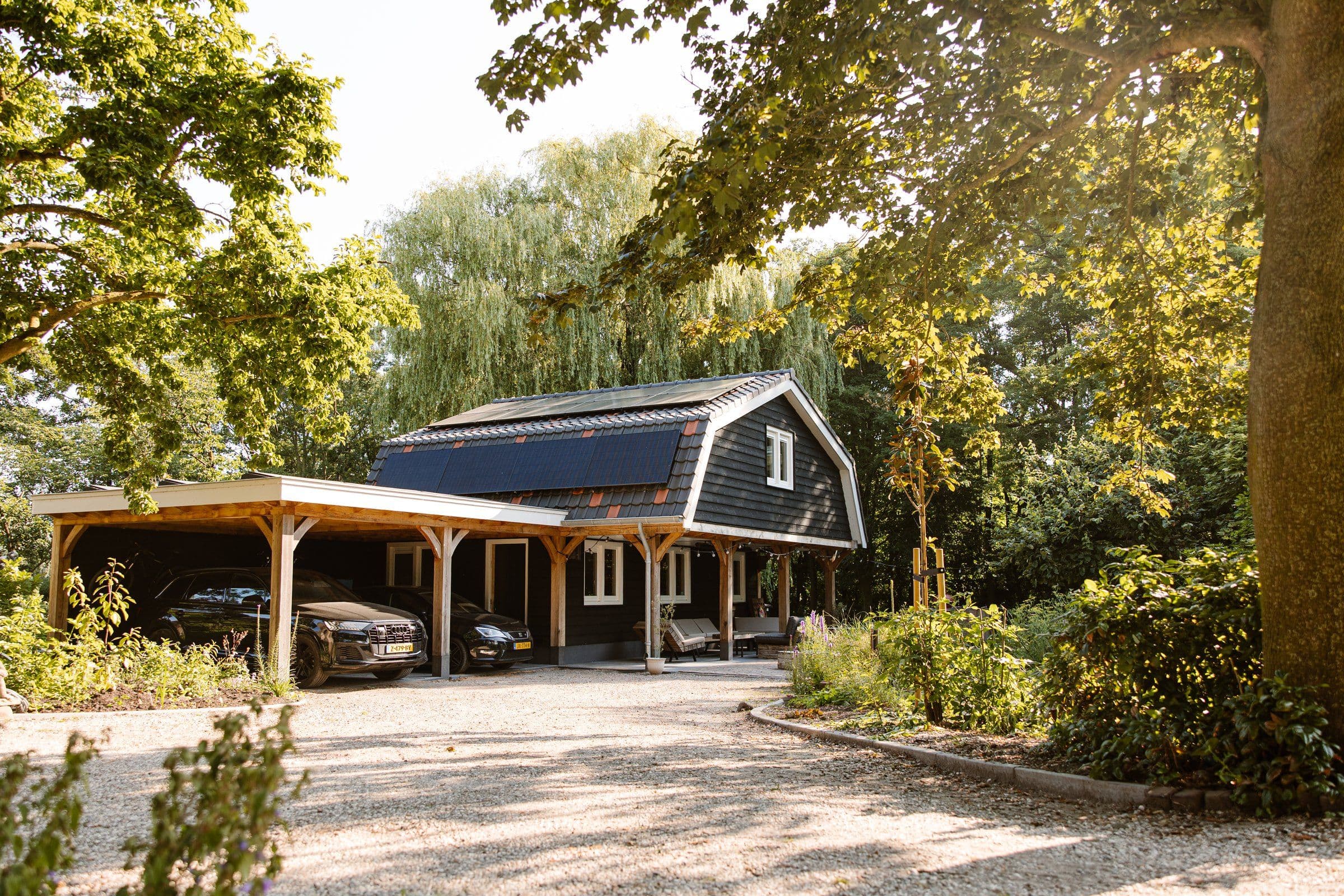 Donker houten huis met zonnepanelen op het dak, overkapping met twee auto's en een groene tuin