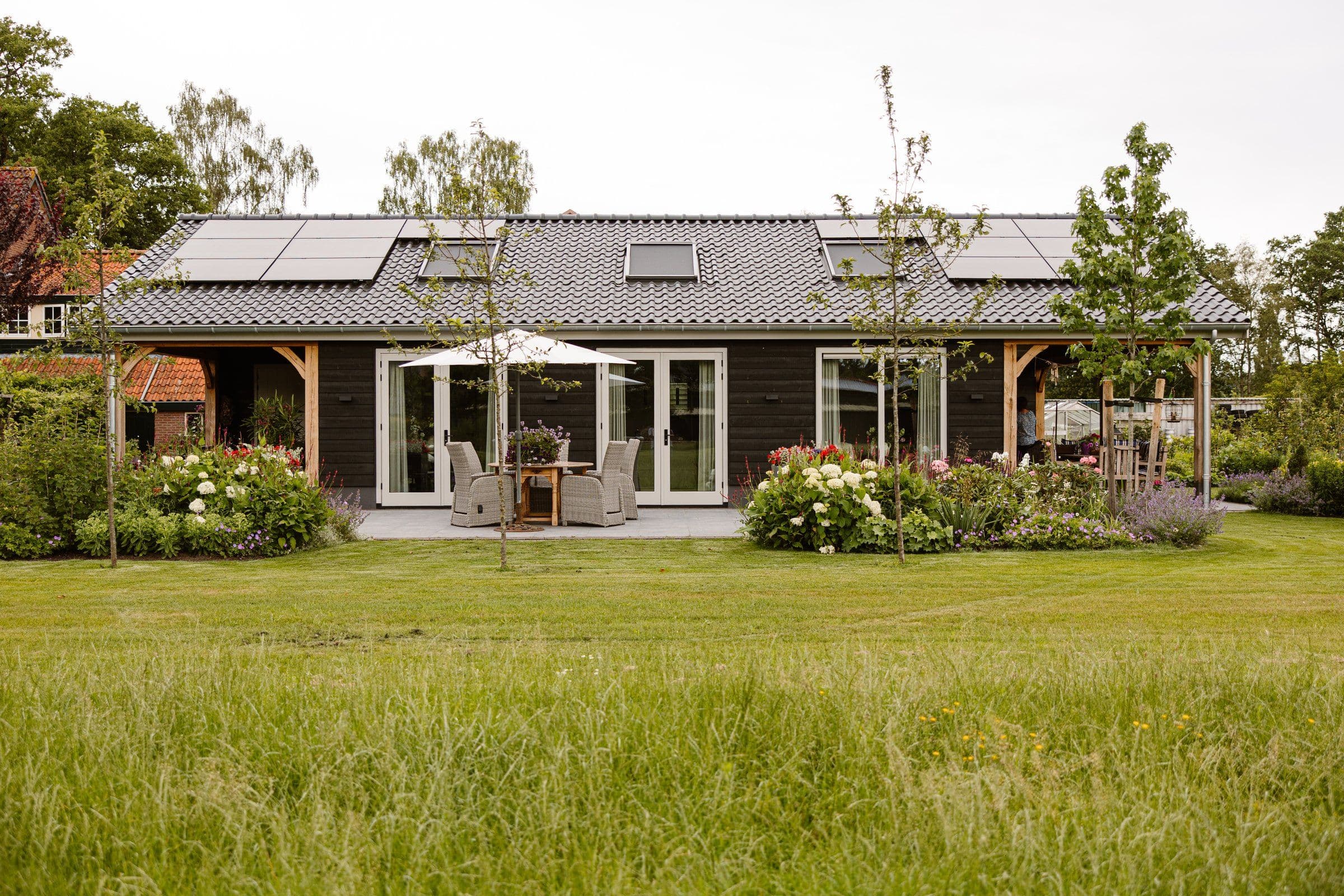 Vrijstaand huis met donkere houten gevel, grijs pannendak met zonnepanelen en dakramen, terras met tuintafel en parasol en voorgelegen bloementuin en grasveld.
