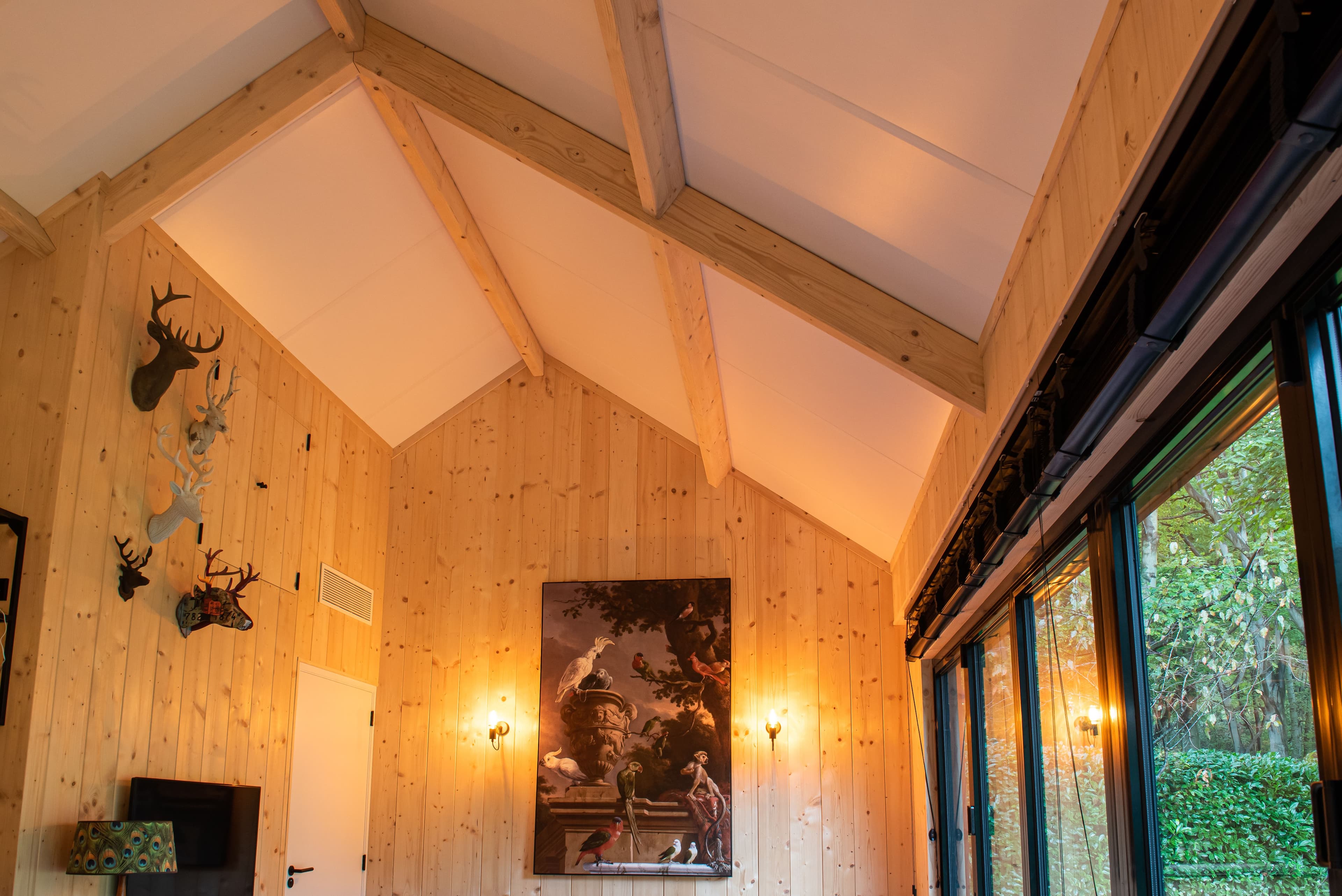 Binnenruimte met houten wanden en plafond, verschillende dierenkoppen aan de muur, schilderij van vogels en grote ramen met uitzicht op natuur.