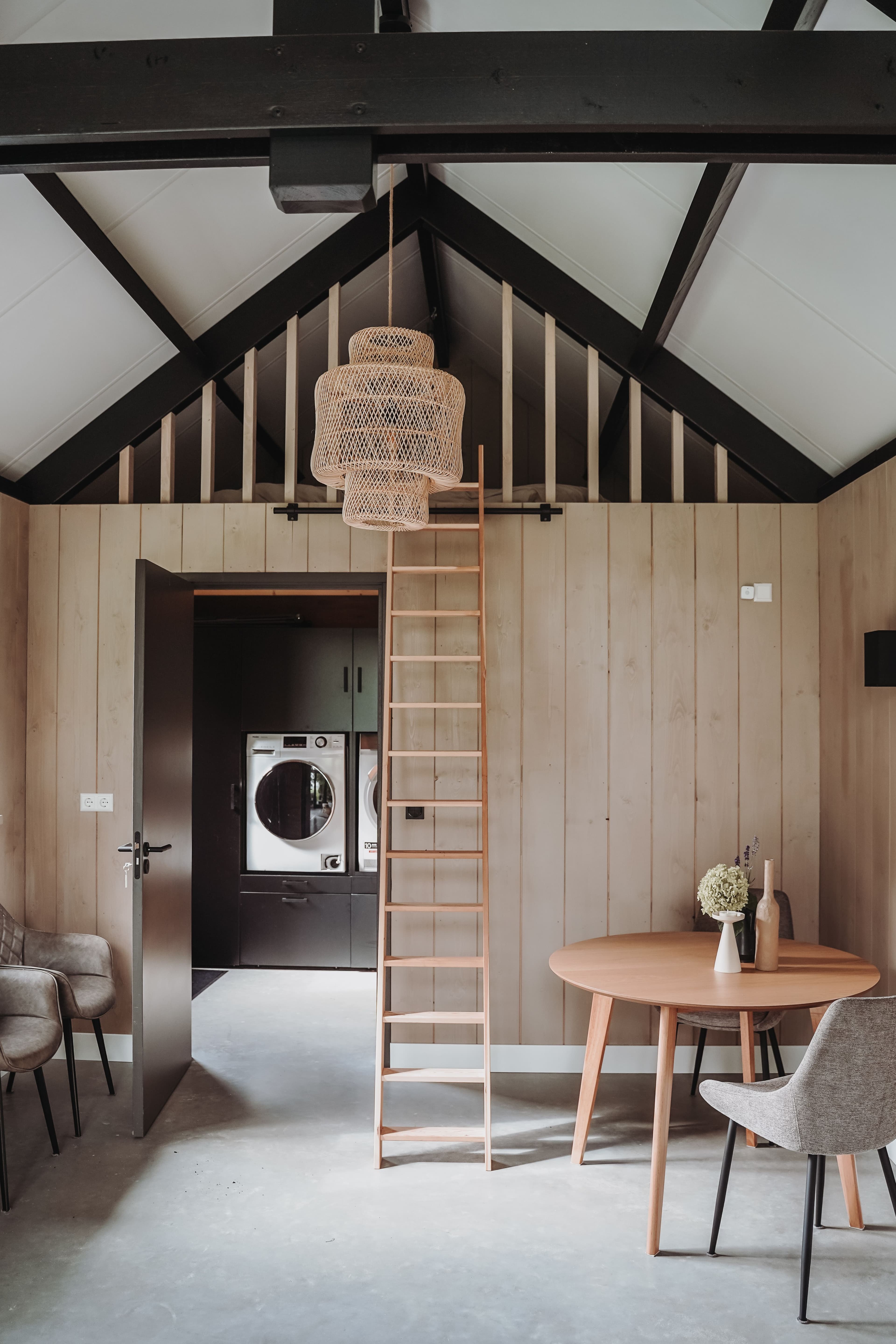 Binneninterieur met houten ladder naar vide, rieten hanglamp en wasmachine zichtbaar in aangrenzende ruimte
