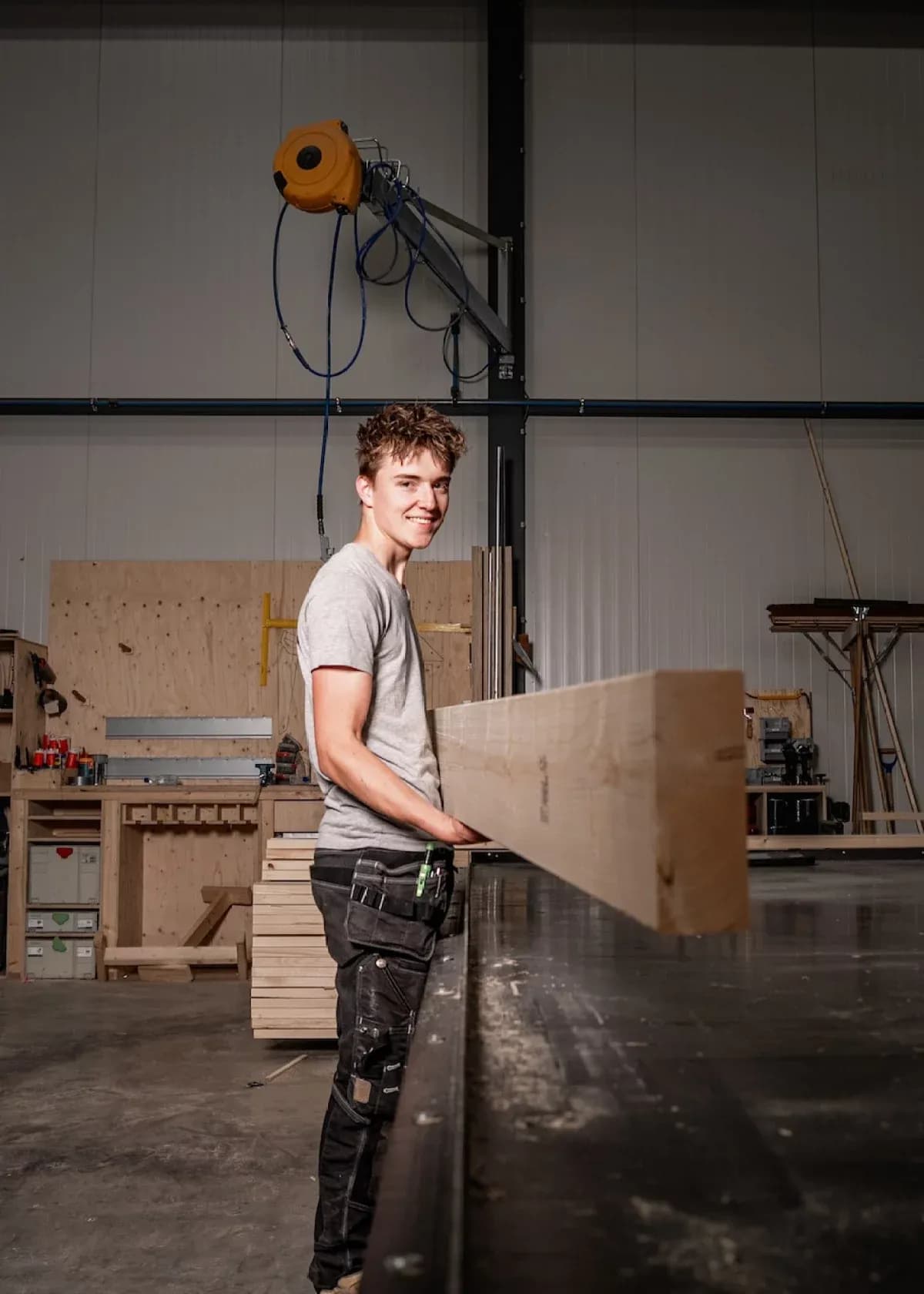 Jonge man met grijs t-shirt en zwarte werkbroek draagt een lange houten balk in een houtwerkplaats