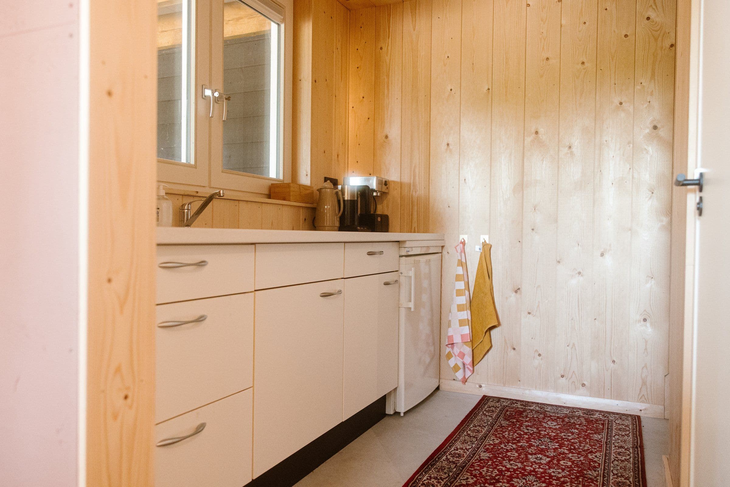Kleine keukenruimte met houten wand, witte kastjes, koffiemachine en kleine koelkast