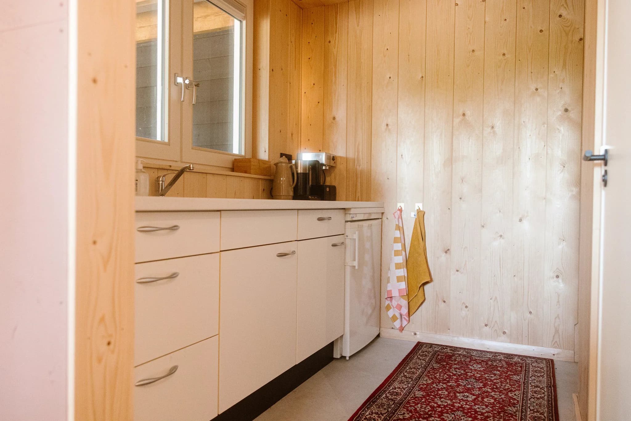 Kleine keukenruimte met houten wand, witte kastjes, koffiemachine en kleine koelkast
