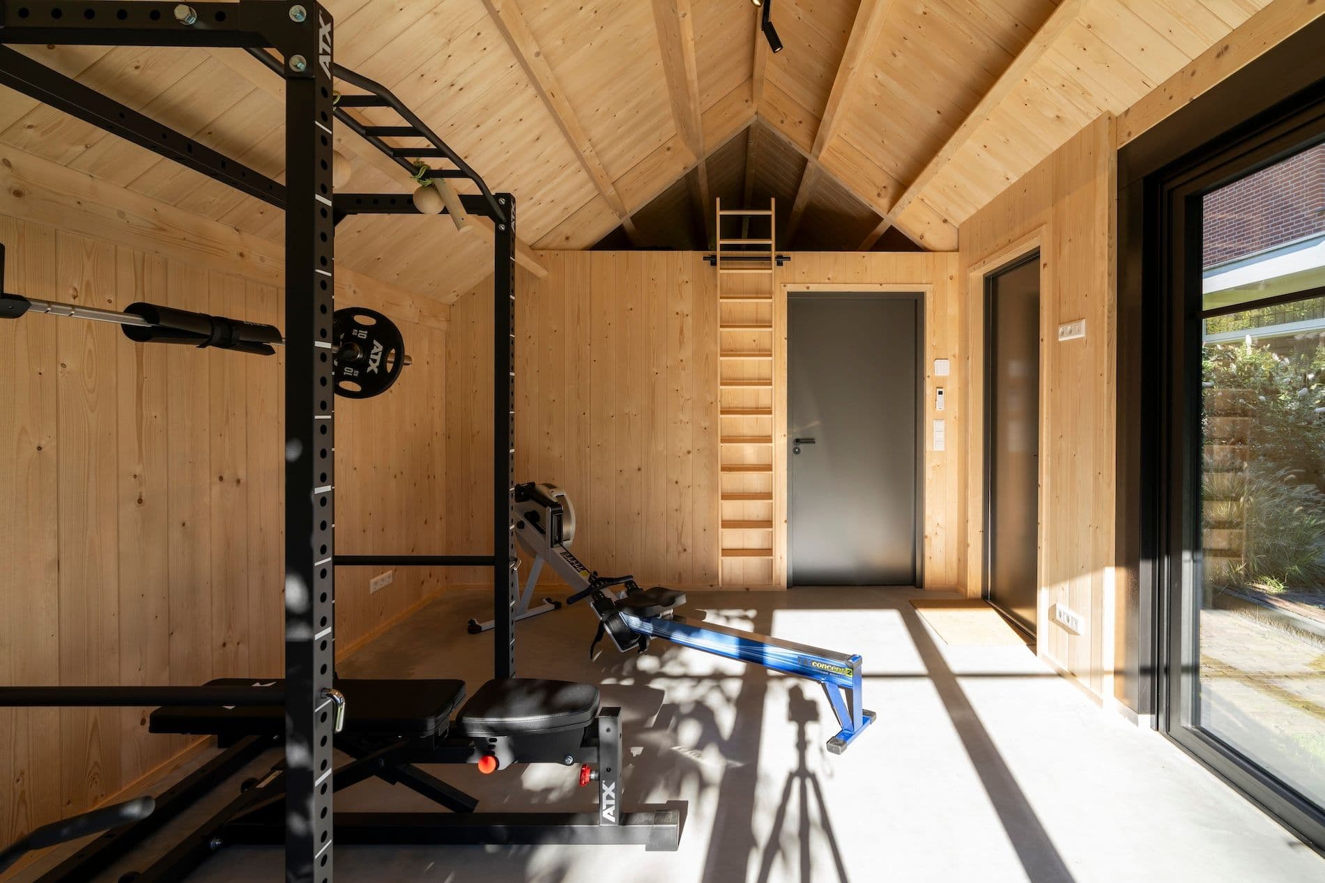 Fitnessruimte met houten wanden en dak, halterbank, roeimachine en grote glazen schuifdeur naar buiten