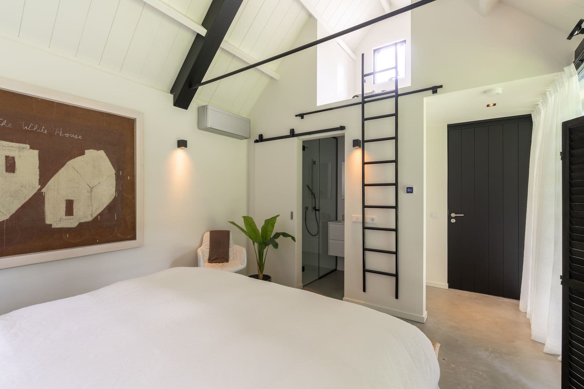 Lichte slaapkamer met schuin plafond, zwarte metalen ladder naar een vide, schuifdeur naar badkamer, zwarte deur en ingelijst doek aan de wand.