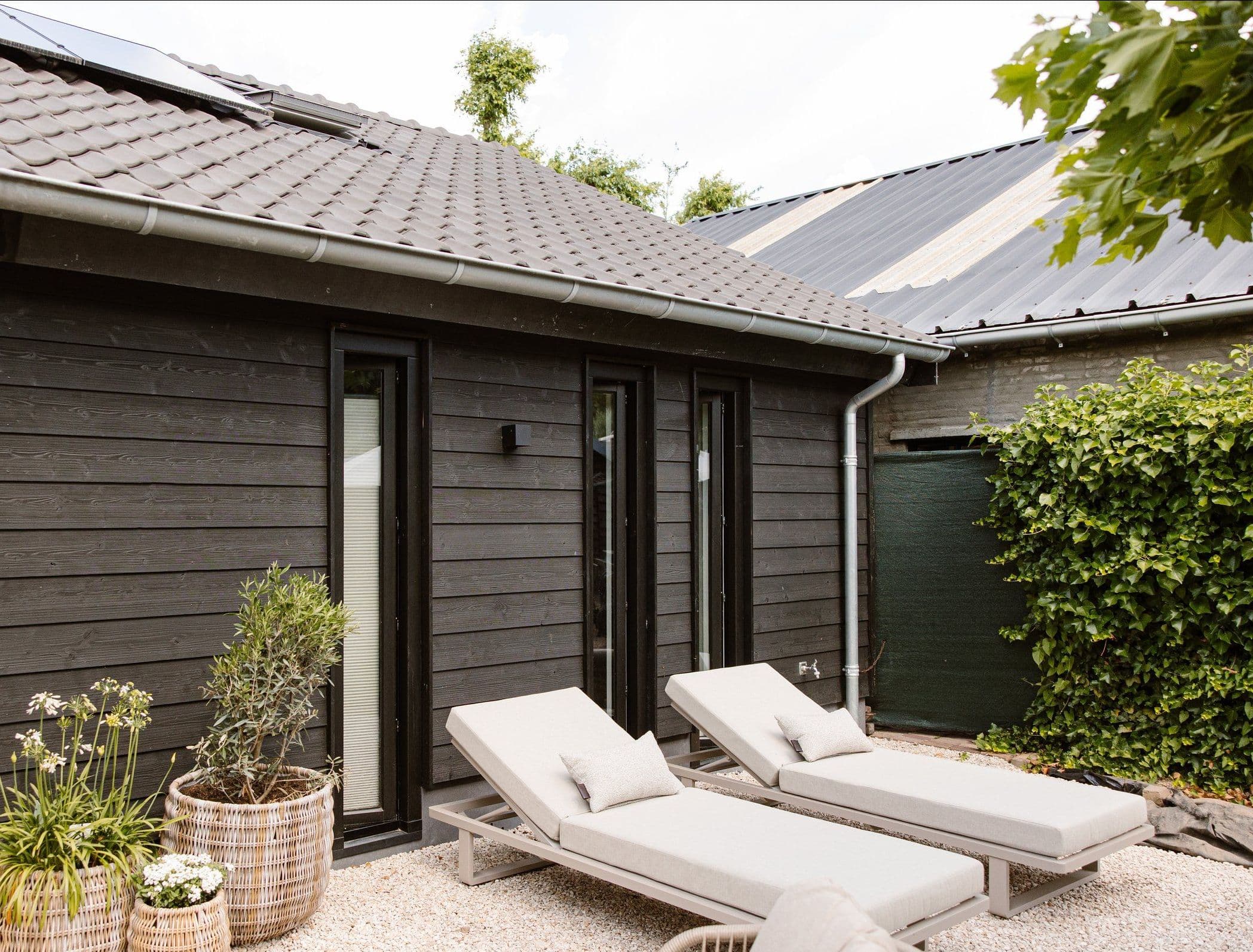 Twee ligbedden met kussens voor een zwart houten huis met smalle deuren, planten in manden en een groene haag