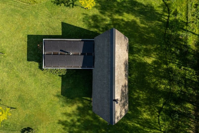 Luchtfoto van een woning met zonnepanelen op het dak omgeven door groen gras en schaduwen van bomen