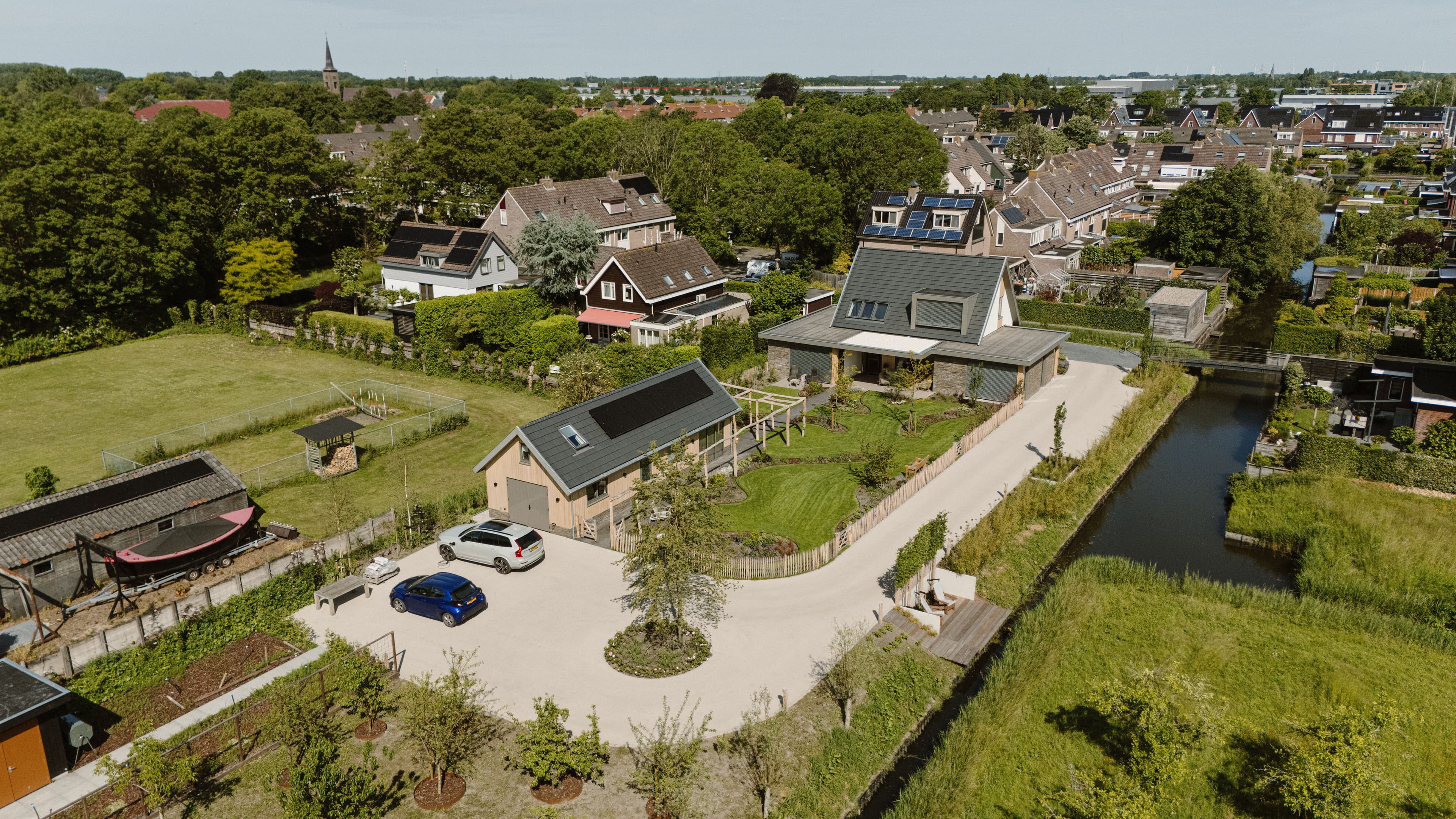 Luchtfoto van een woonwijk met huizen, tuinen, een smalle watergang, geparkeerde auto's en zonnepanelen op meerdere daken