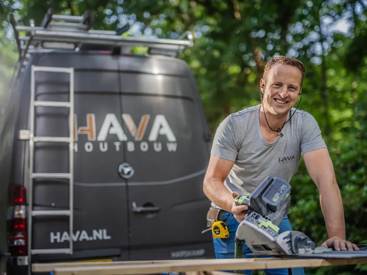 Man in grijs shirt zaagt hout met elektrische zaag, een bus met HAVA HOUTBOUW logo op de achtergrond