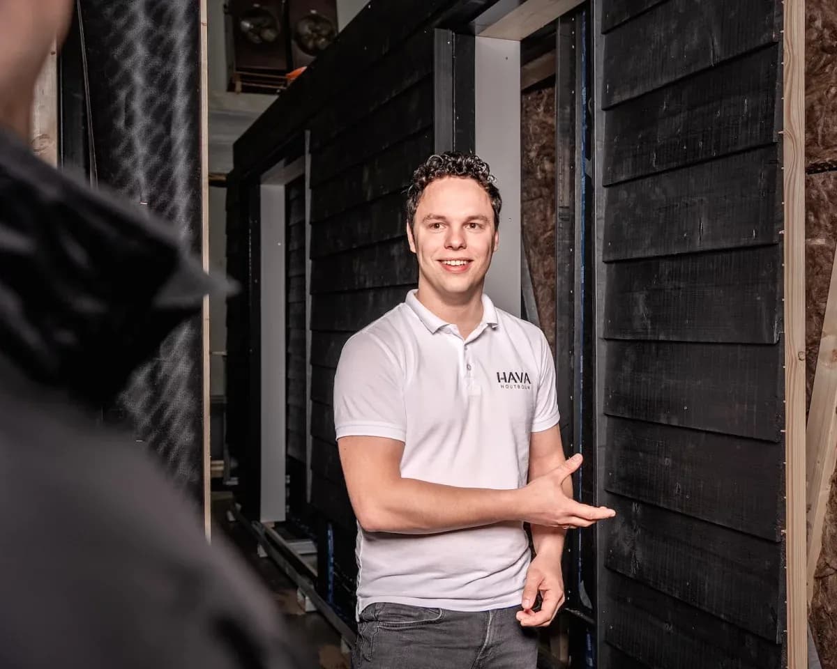 Man in wit poloshirt naast zwarte houten wand die iets uitlegt