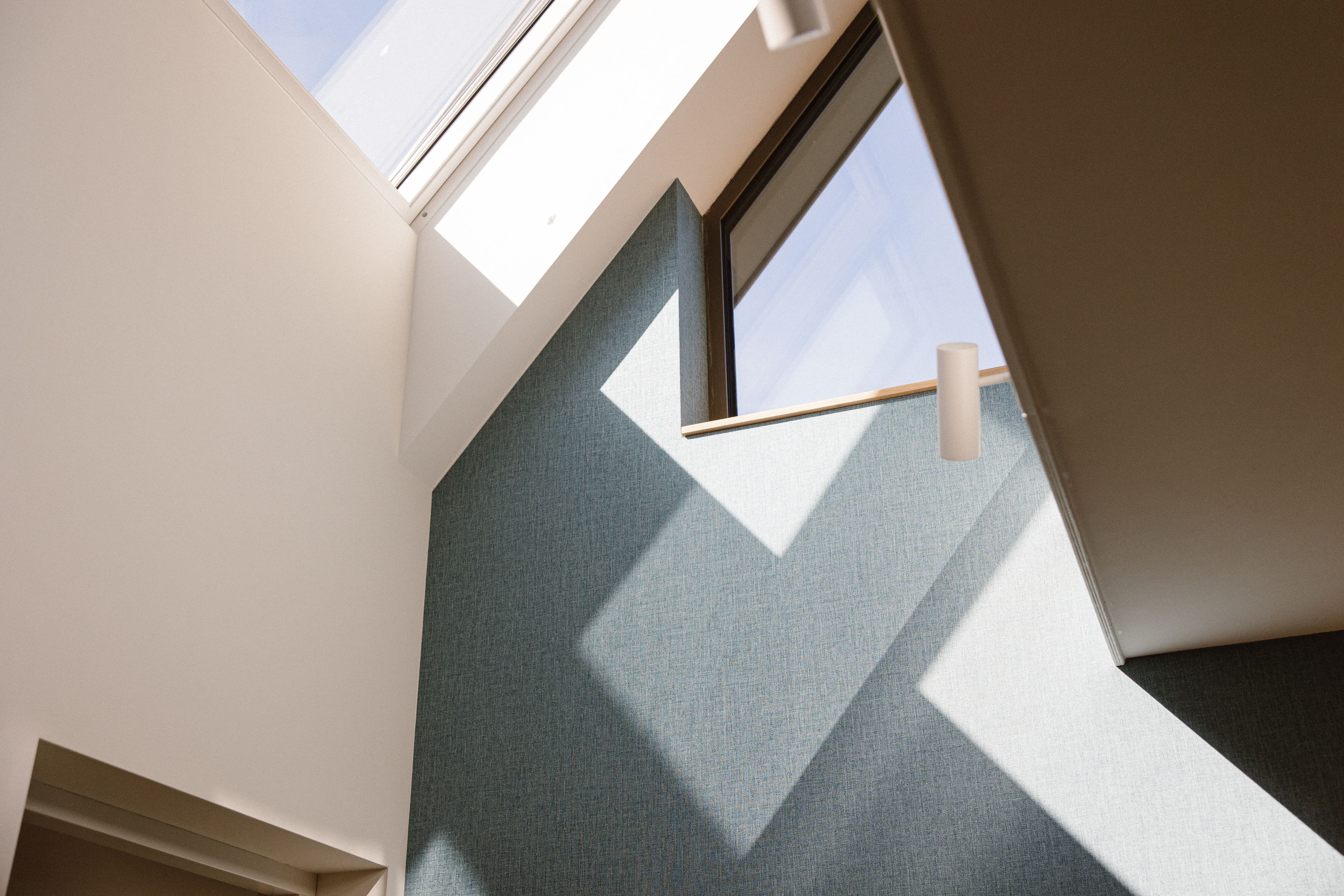 Binnenzicht van een modern gebouw met hoge plafonds en grote ramen waar licht en schaduwen op een blauwe muur vallen.