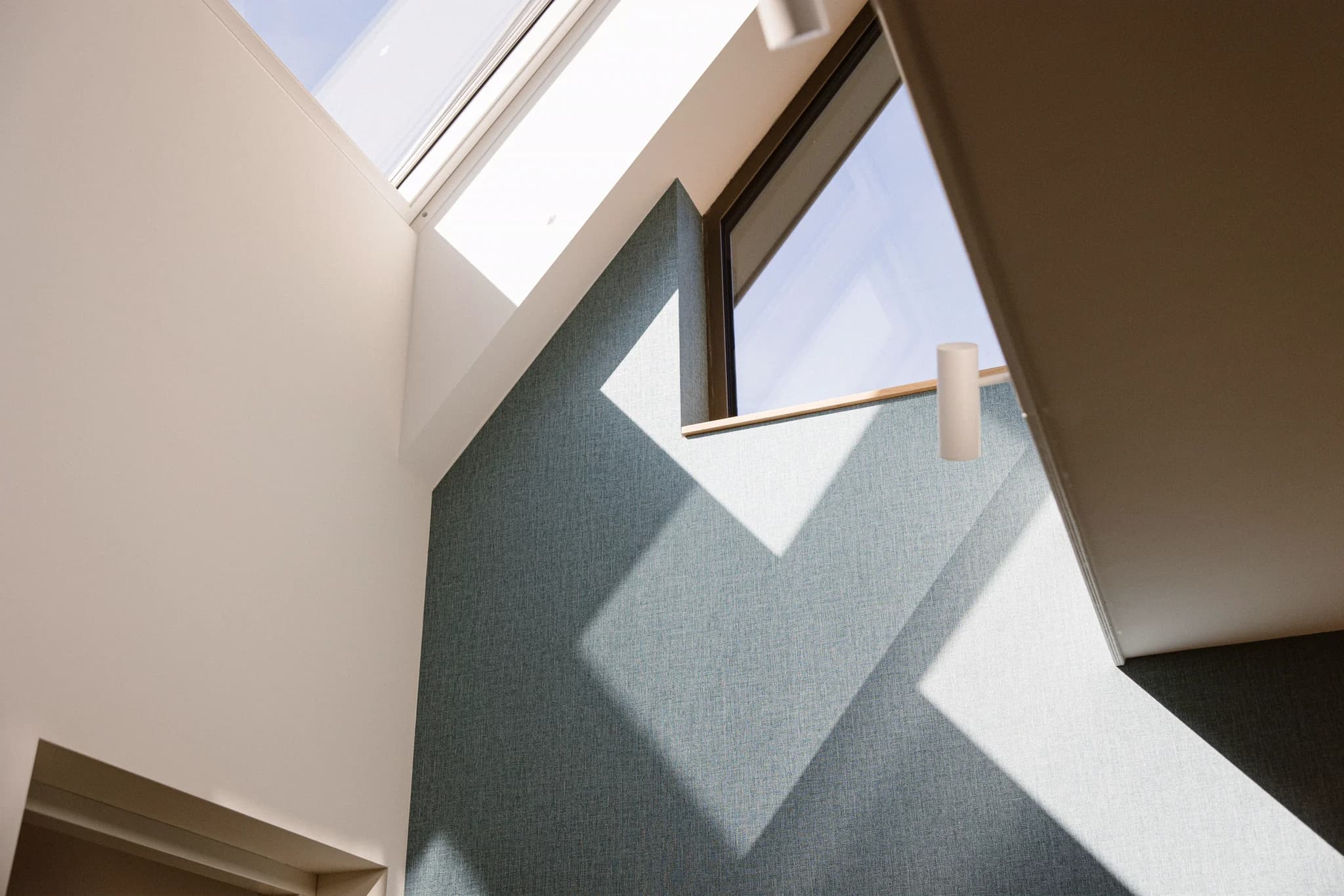Binnenzicht van een modern gebouw met hoge plafonds en grote ramen waar licht en schaduwen op een blauwe muur vallen.