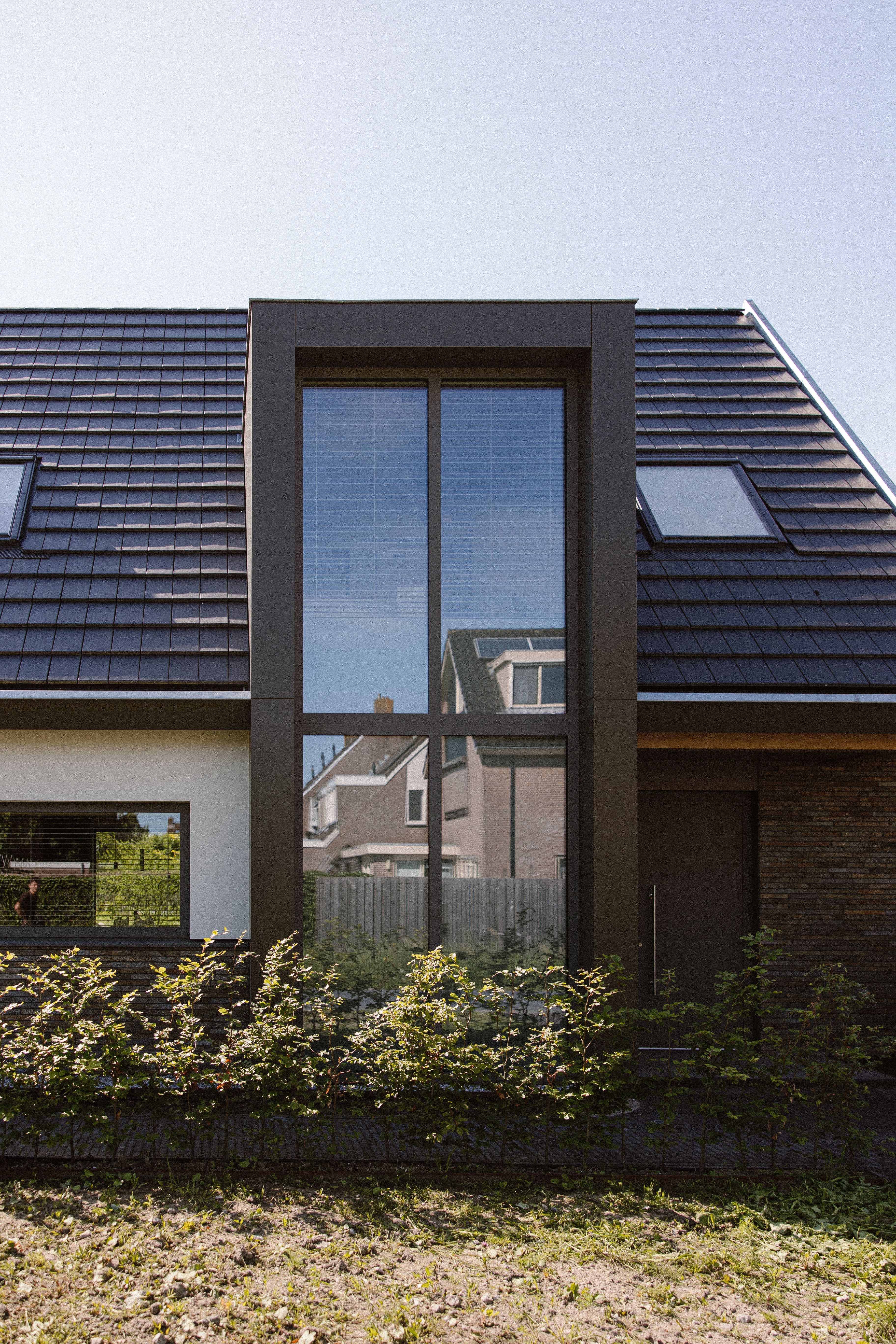 Modern huis met een groot uitbouwraam in het midden, dakramen en een heg aan de voorzijde.