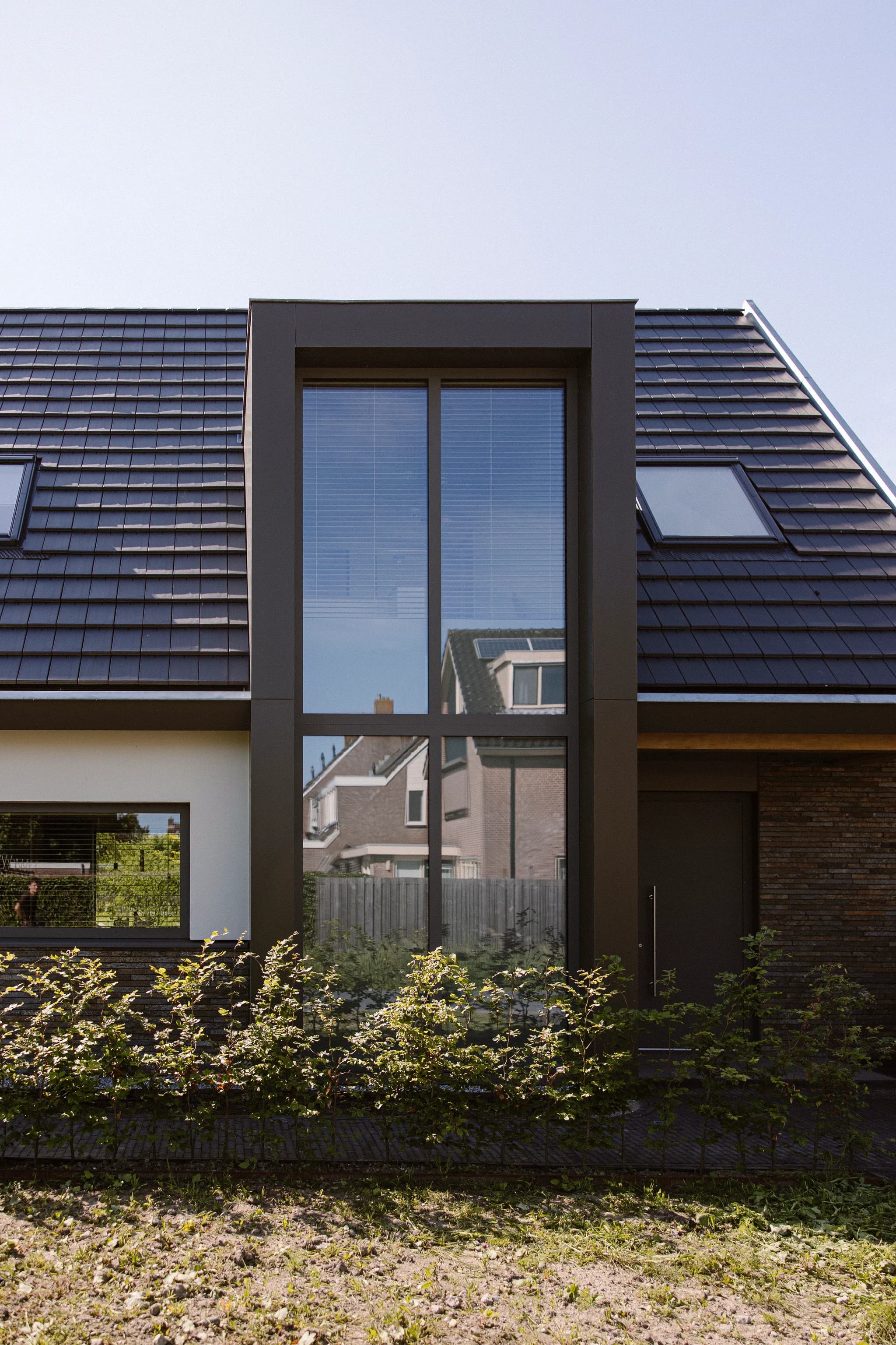 Modern huis met een groot uitbouwraam in het midden, dakramen en een heg aan de voorzijde.