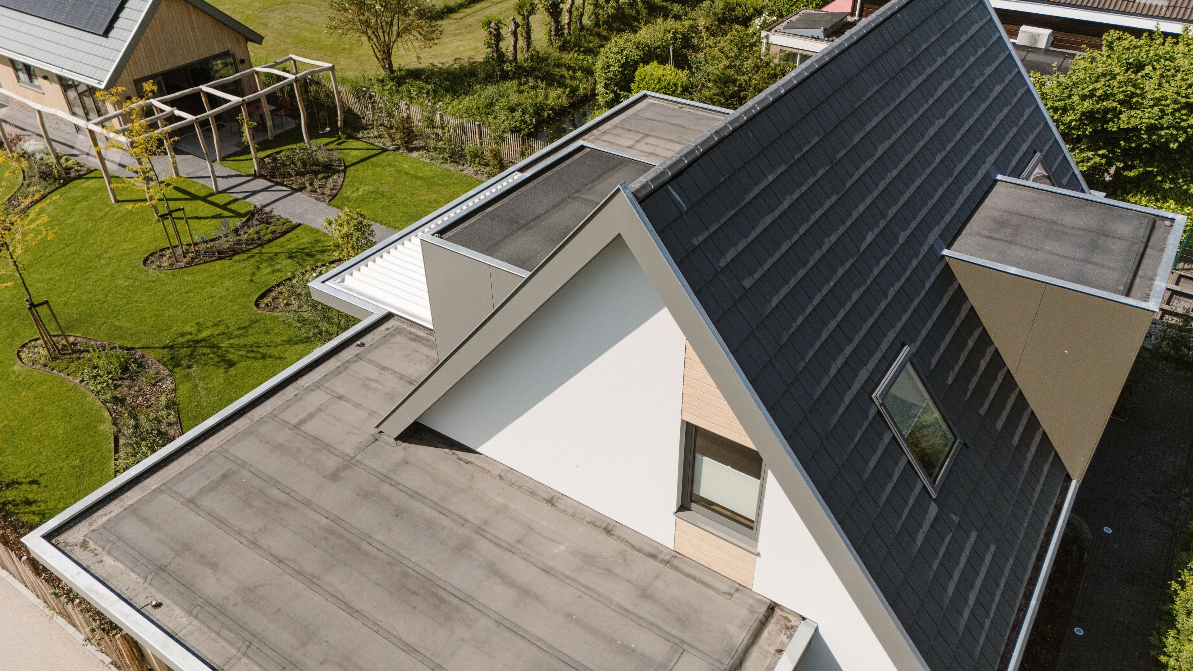 Luchtfoto van een huis met plat dak en schuindak met donkere pannen, omgeven door een tuin met gras en bomen.