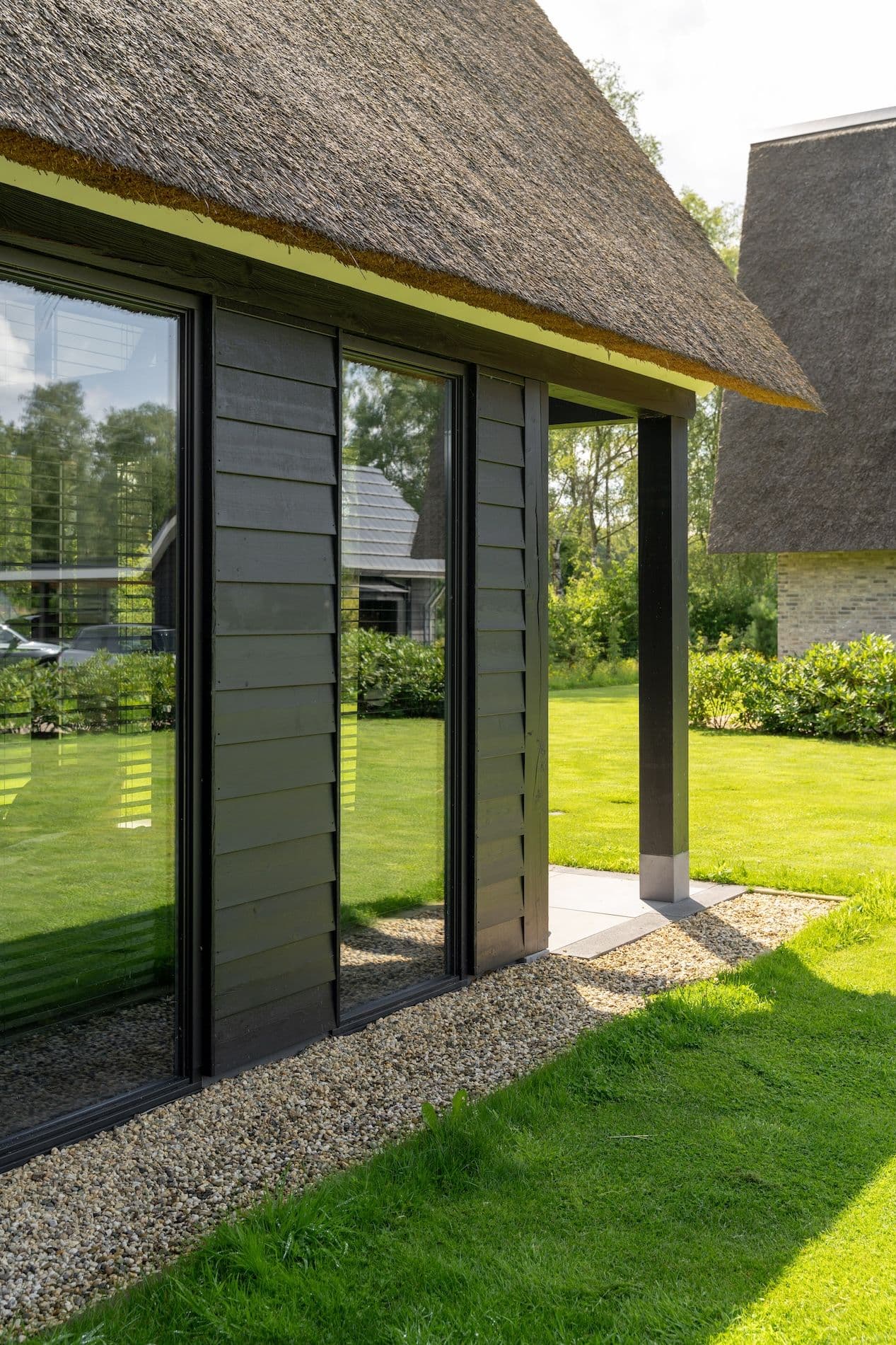 Zijaanzicht van een modern huis met zwarte houten gevelbekleding, glas en een rieten dak aan een groene tuin met gazon