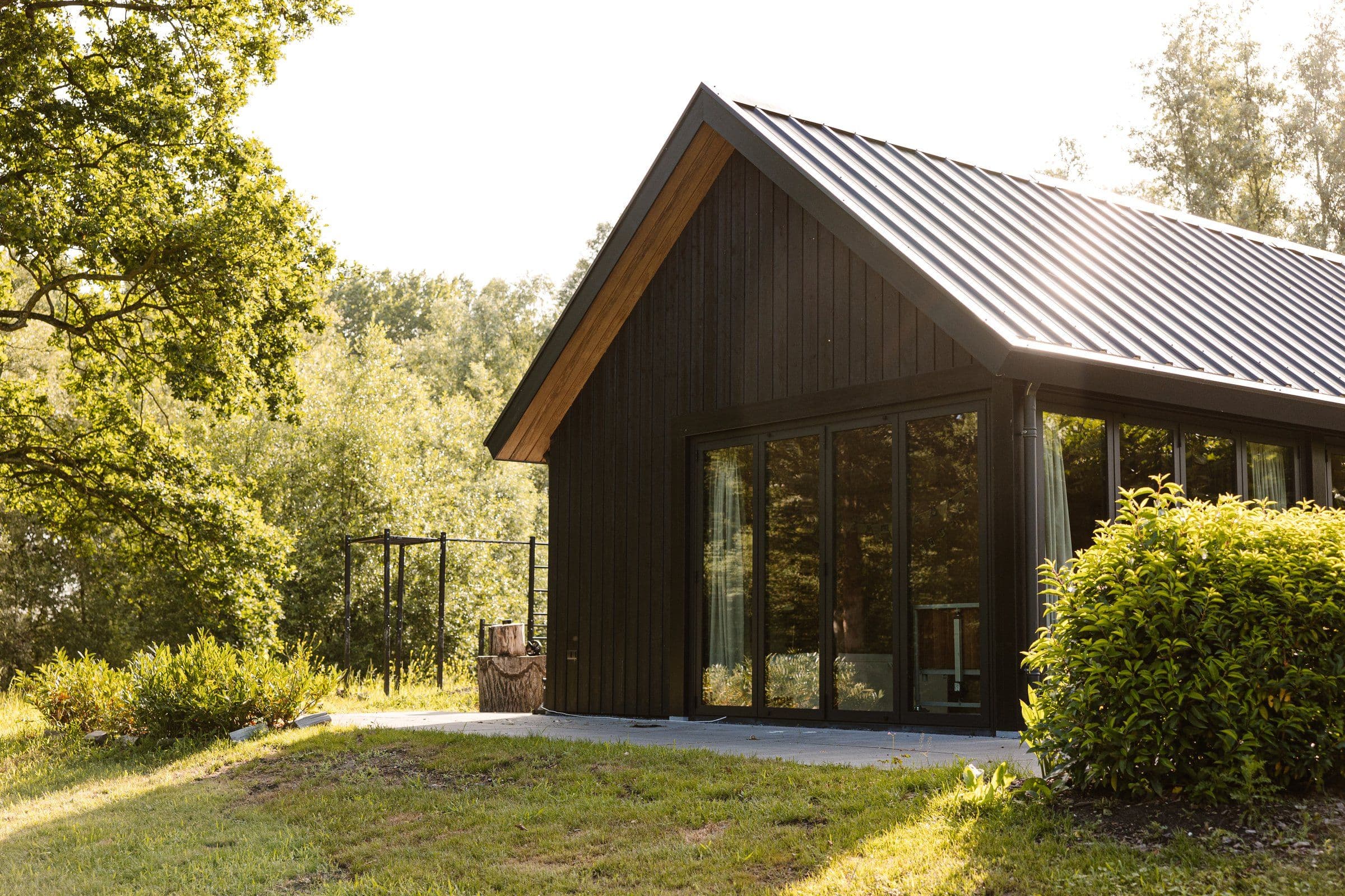 Zijkant van een modern zwart houten huis met grote ramen en groen gras en struiken rondom