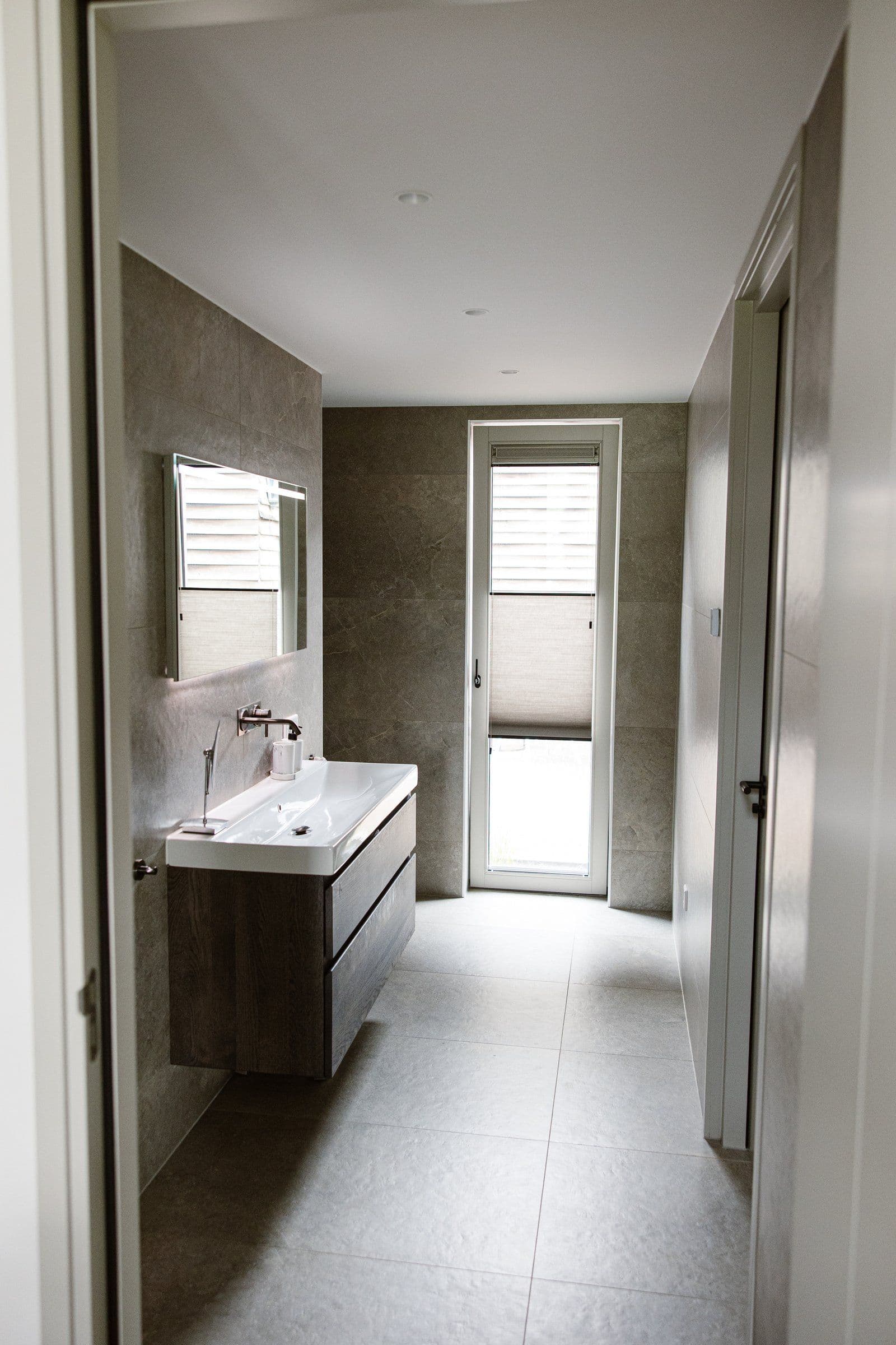 Interieurfoto van een moderne badkamer met een zwevend wastafelmeubel, grote spiegel en een smal raam met stofrolgordijn.