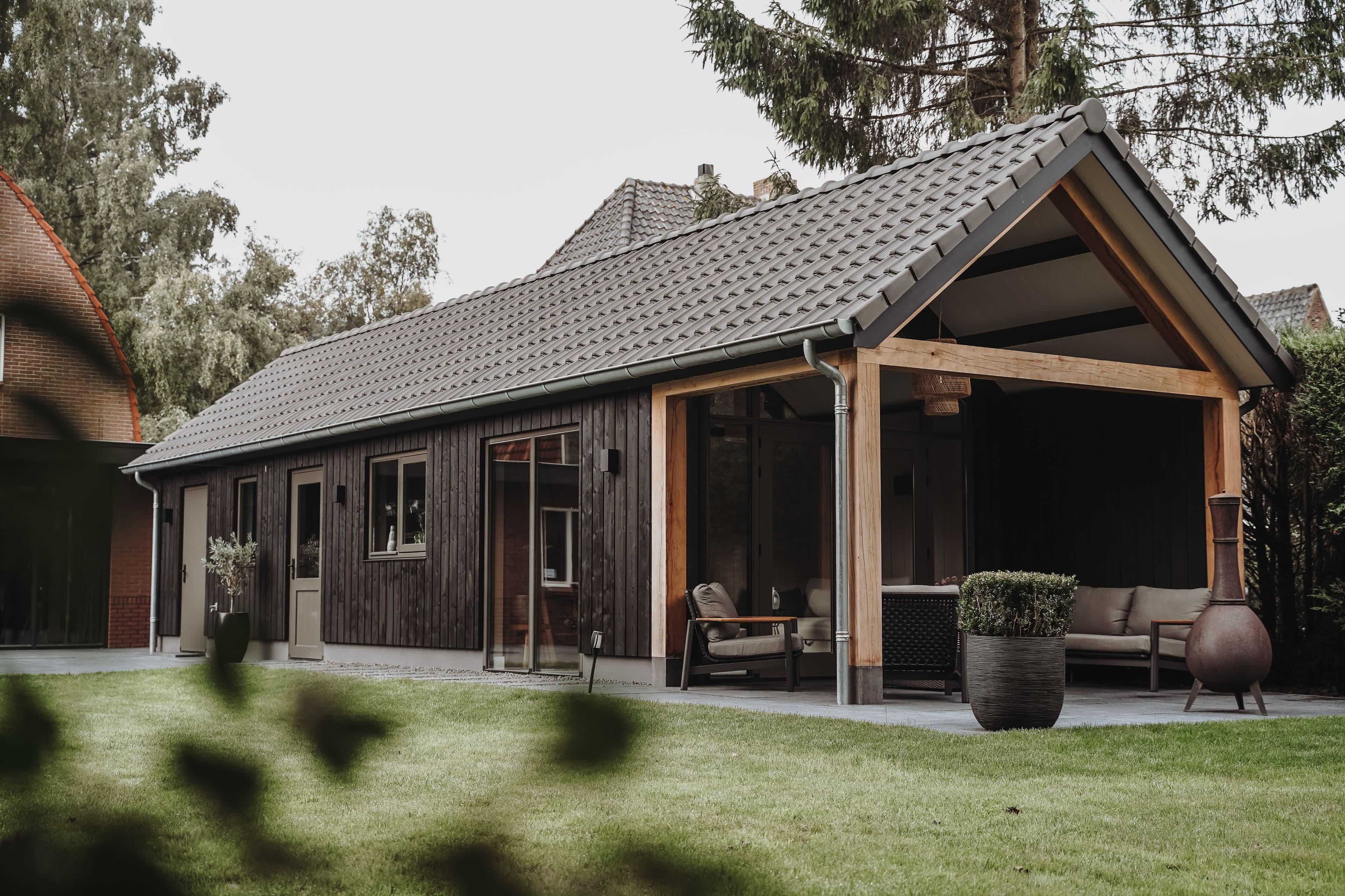 Moderne bungalow met donkere houten gevelbekleding en een overdekt terras met stoelen en plantenbak, gelegen aan een groene tuin
