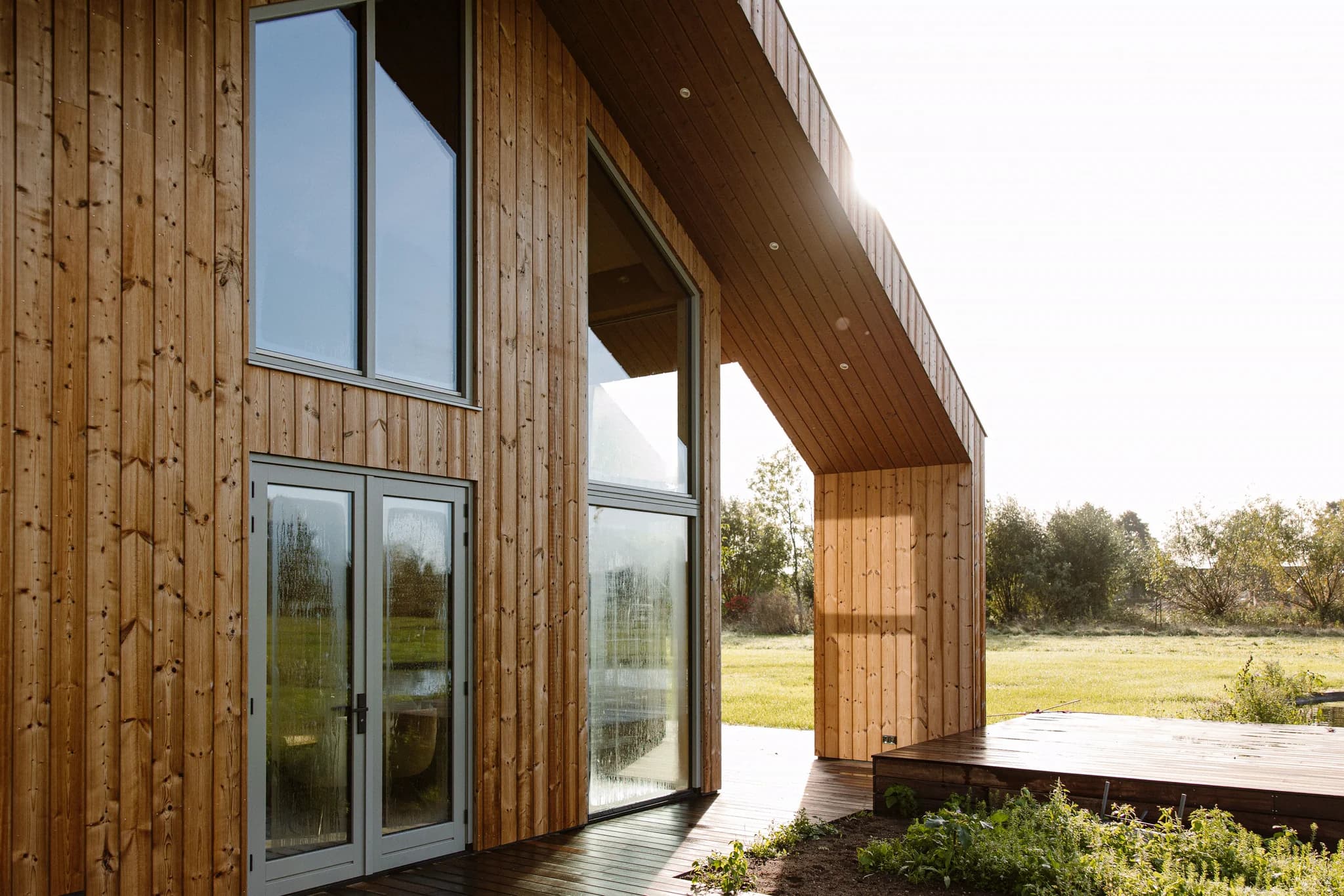 Houten gevel van een modern huis met grote ramen en een houten terras met uitzicht op een groene tuin