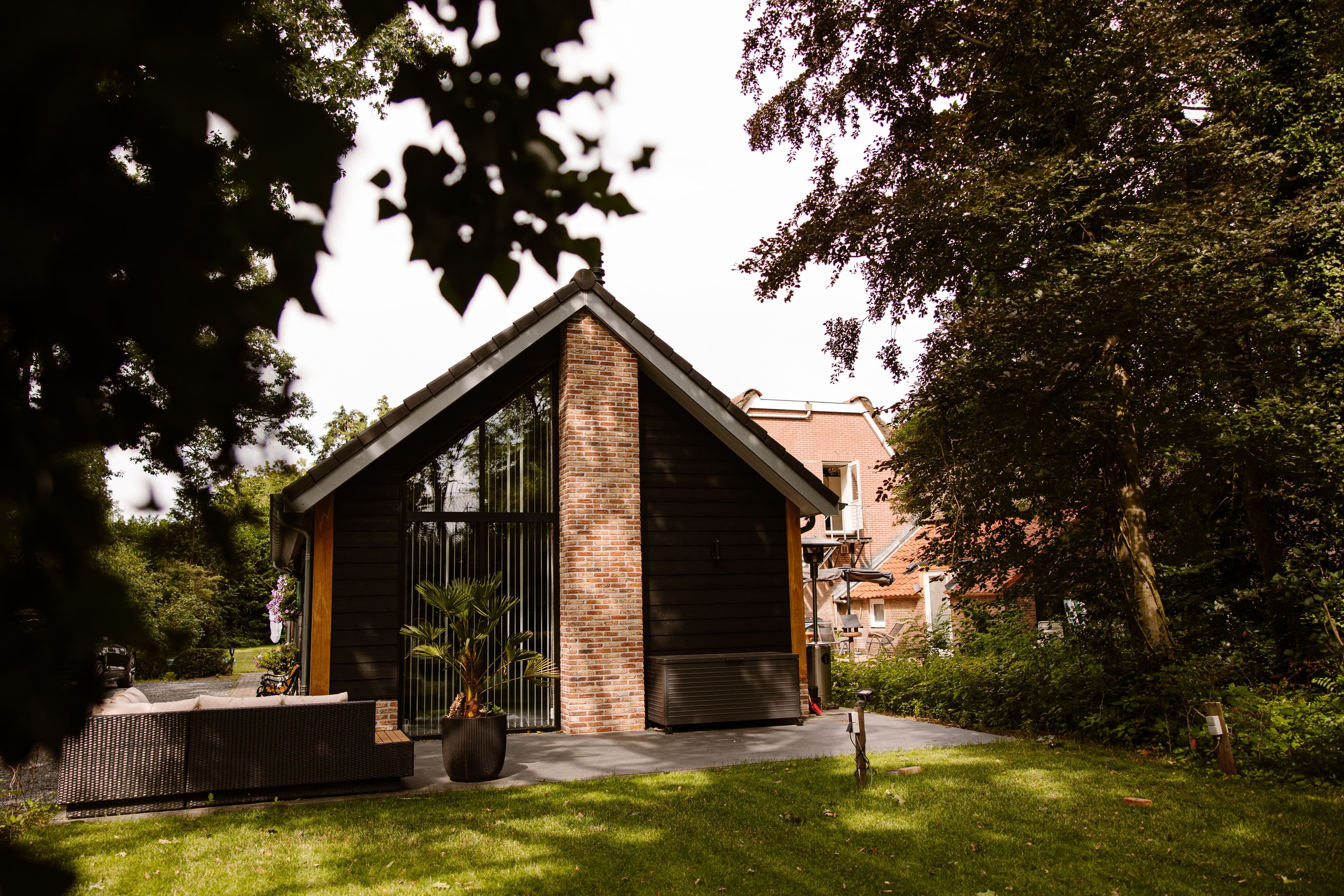 Moderne houten schuur met grote ramen en bakstenen schoorsteen gelegen in een tuin met groen gras en bomen