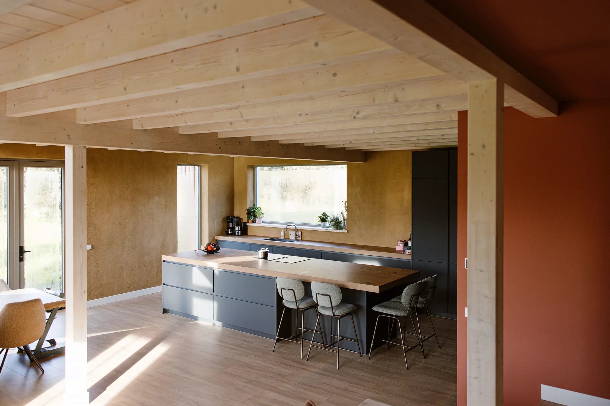 Moderne keuken met donkergrijze kasten, houten werkblad en vier barkrukken in een lichte ruimte met houten balkenplafond
