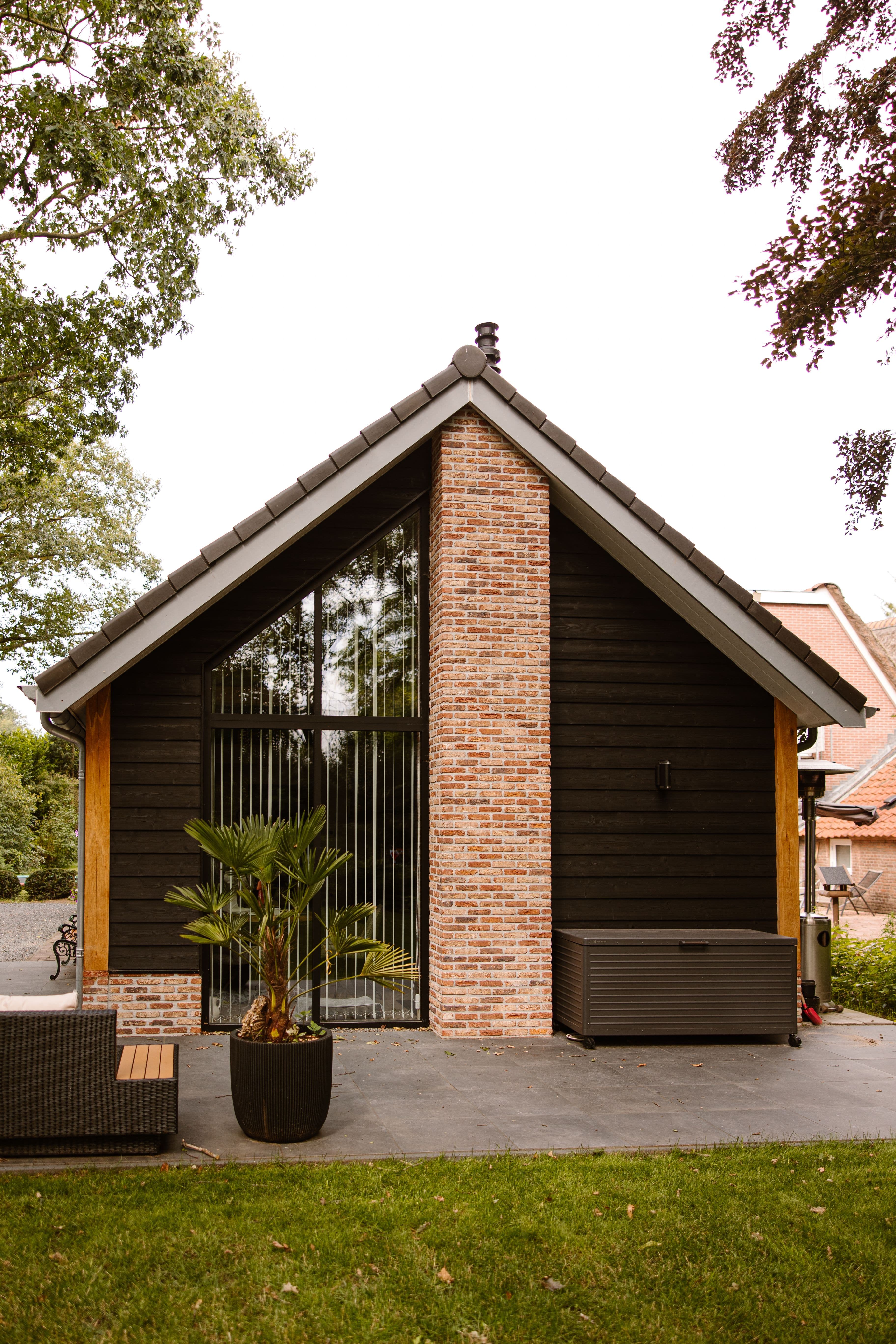 Achterzijde van een schuur met zwart houten gevel, gemetselde schoorsteen, grote glazen deur en een plantenbak met palmplant op een betegeld terras