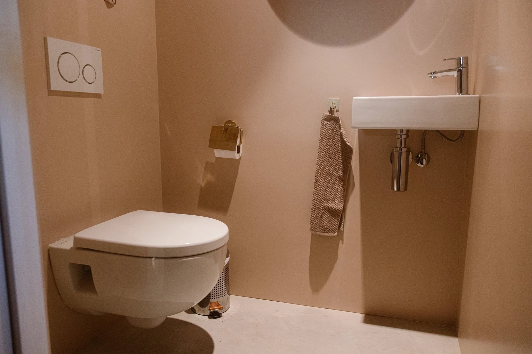 Moderne toiletruimte met hangend wit toilet, klein witte wastafel, beige handdoek aan de muur en een prullenbak op de vloer
