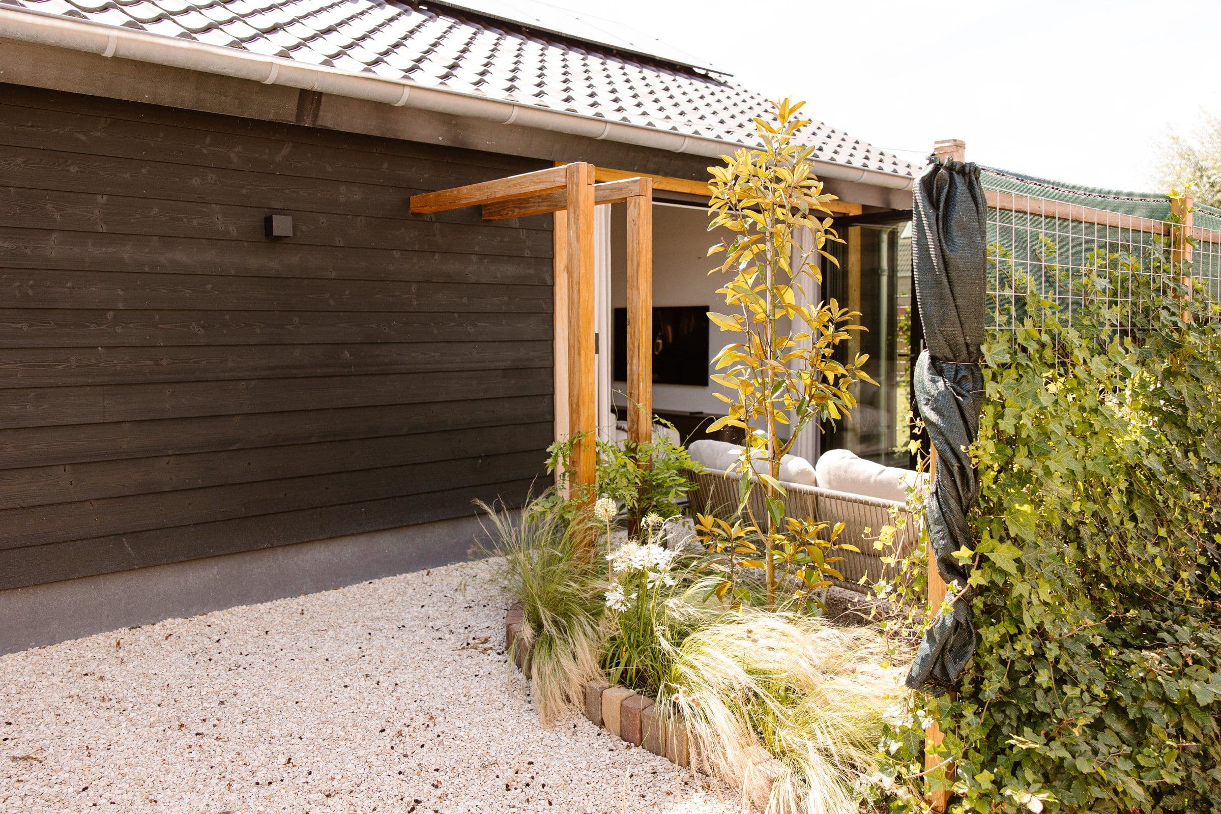 Tuin met grind, een houten wand, pergola en diverse groene planten naast een groot raam met zicht op binnenruimte