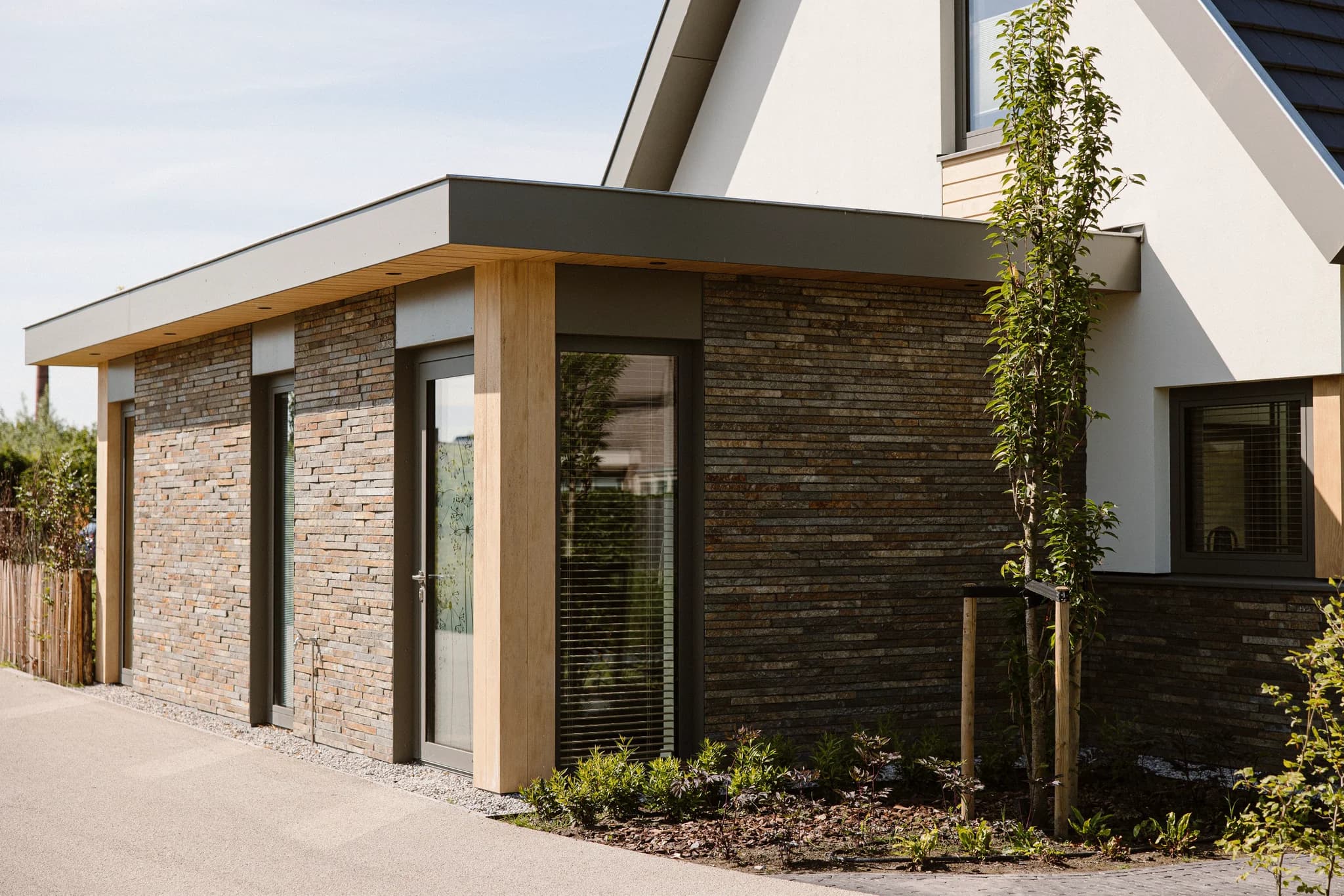 Uitbouw van een huis met stenen gevel en grote ramen naast een jonge boom en een tuin