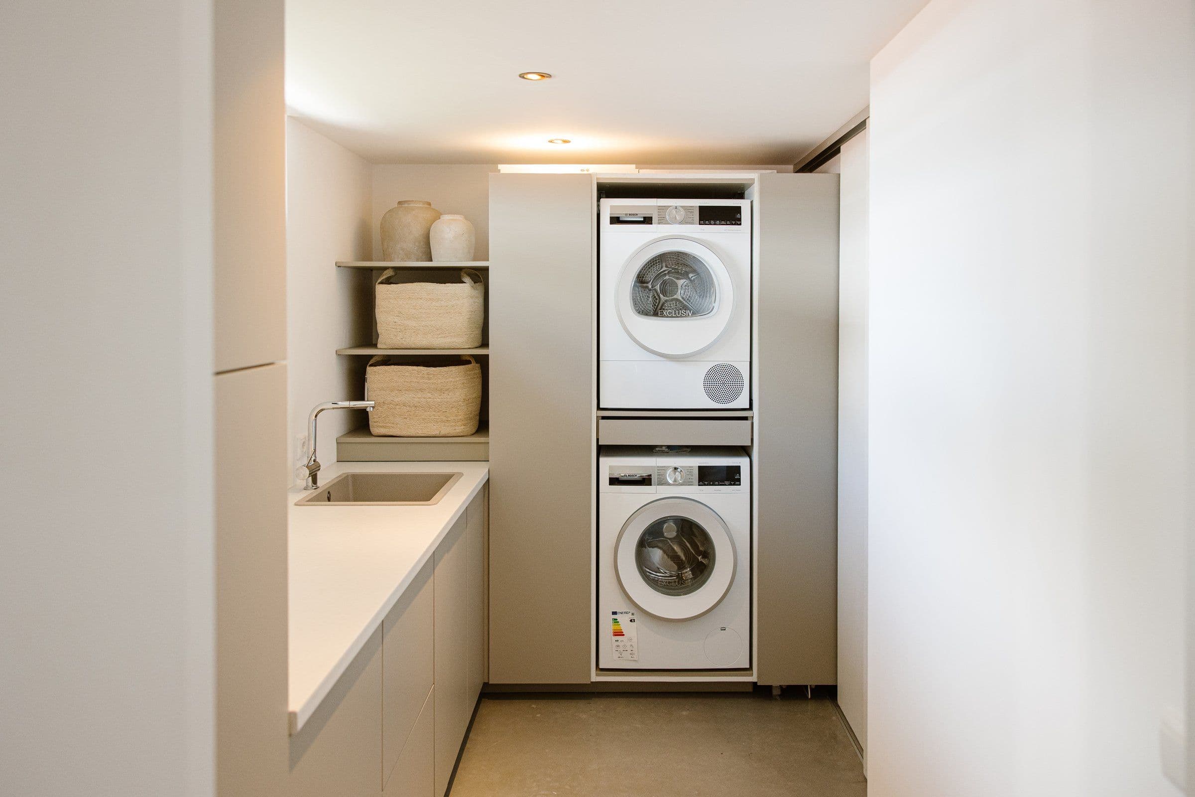 Witte gestapelde wasmachine en droger in een strakke wasruimte met opbergmanden en een gootsteen